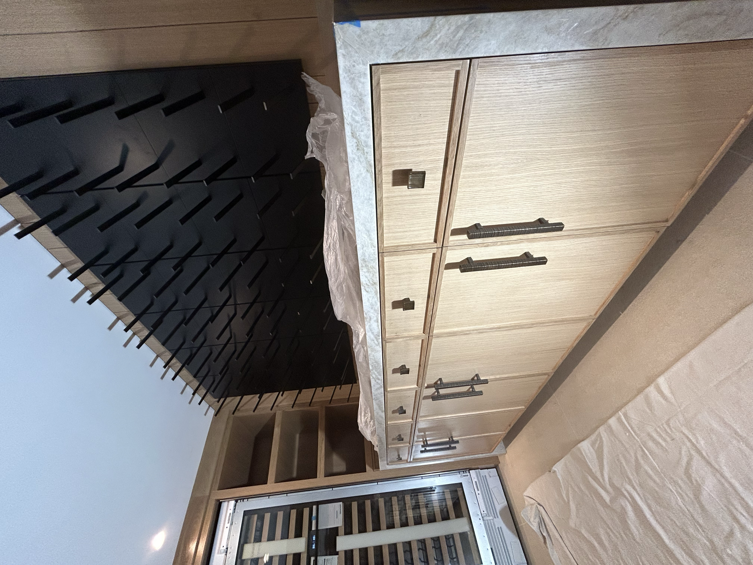 Interior view of a kitchen with wooden cabinets, black hardware, a countertop, a built-in wine cooler, and an open shelf, with part of a covered surface and a black wine rack visible. 