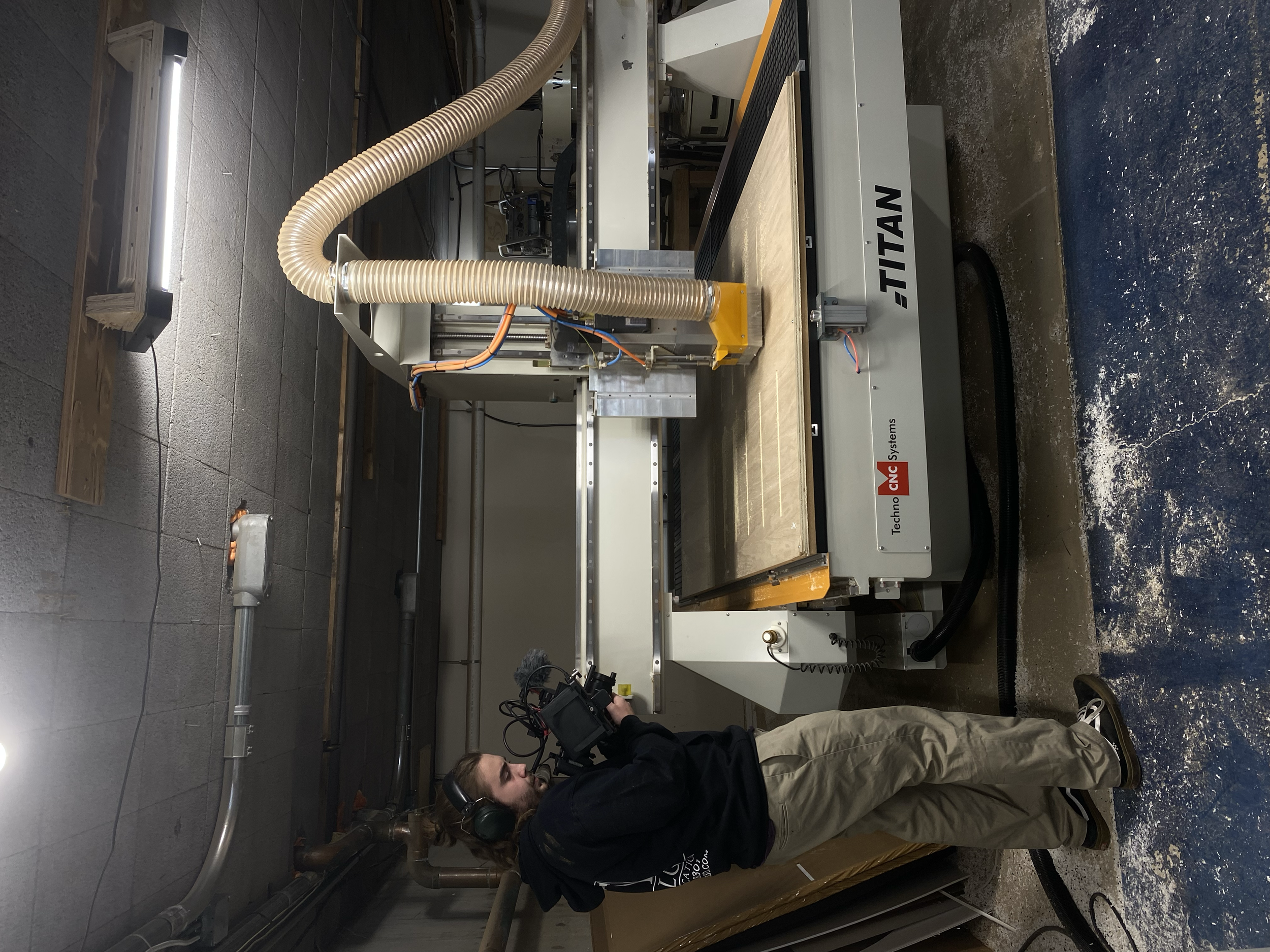 A person operating a CNC machine in a workshop with a gray wall and pipes, with chestnut-colored wood on the CNC machine table.