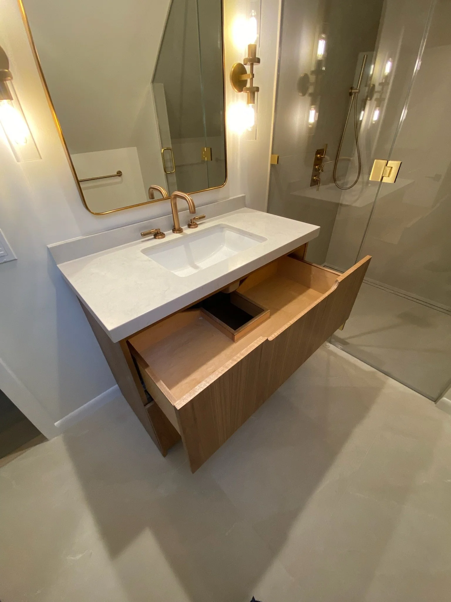 Modern bathroom with a wooden vanity featuring an open drawer, a white countertop with an integrated sink, gold fixtures, a large mirror, wall-mounted gold light fixtures, and a glass-enclosed shower area with a gold showerhead and controls.