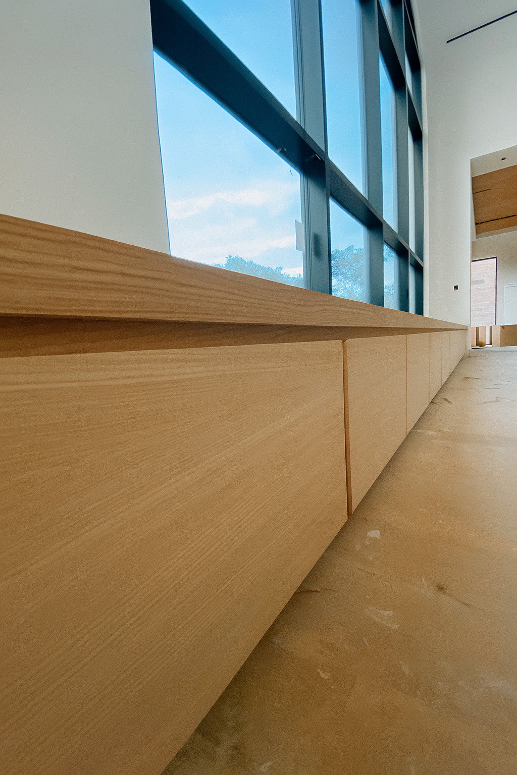 Interior of a modern building with a low wooden cabinet, large floor-to-ceiling windows showing a partly cloudy sky, and a beige marble floor. cabinet renovation. woodwork.