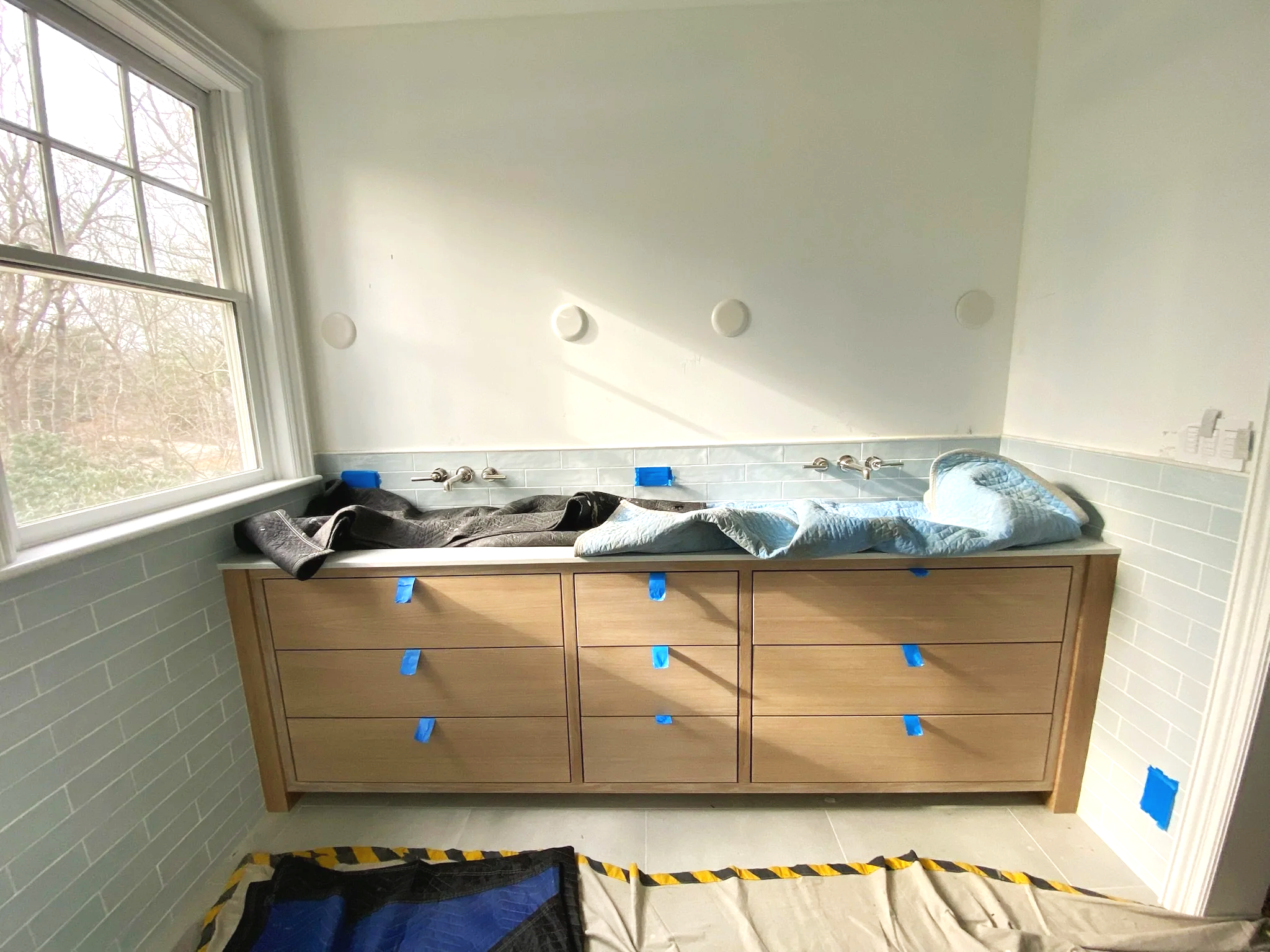 Bathroom with a wooden vanity under a window, with a bathtub filled with water and towels, and masking tape on drawers and wall.