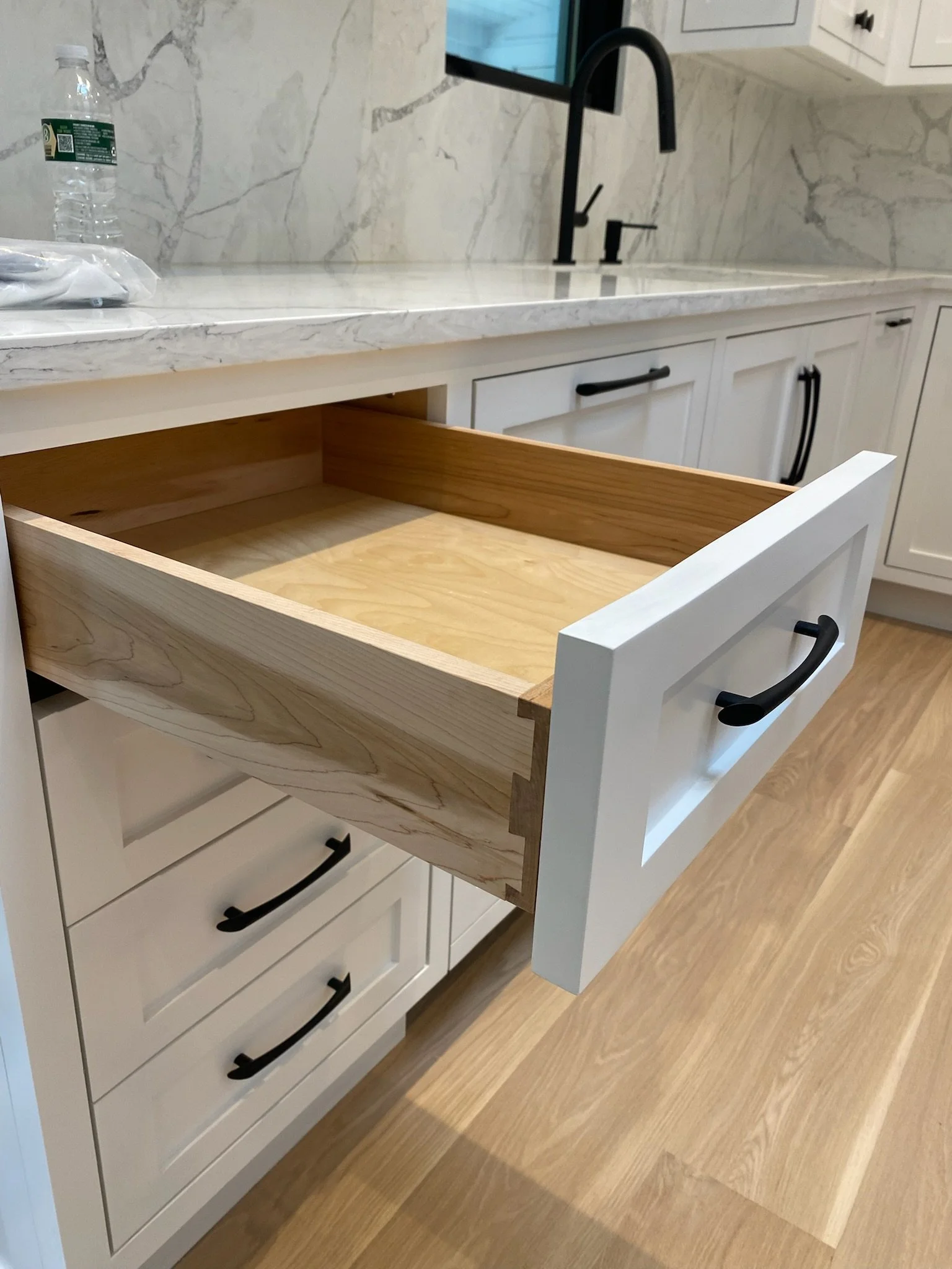 A kitchen drawer with a white front, black handle, and unfinished wooden interior partially pulled out from white cabinets under a marble countertop.