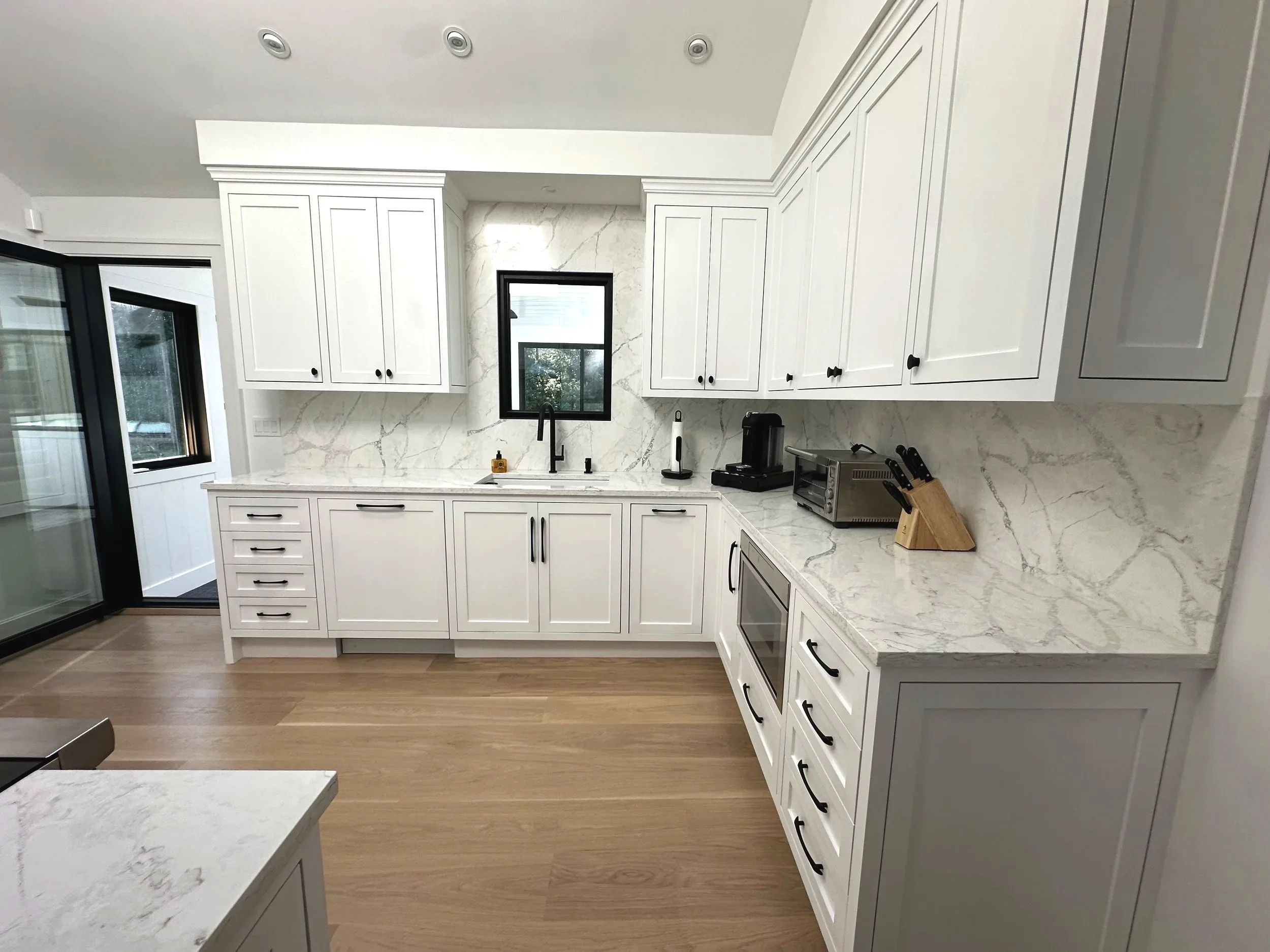Modern kitchen with white cabinets, marble countertops and backsplash, black handles, a small window above the sink, and wooden flooring.