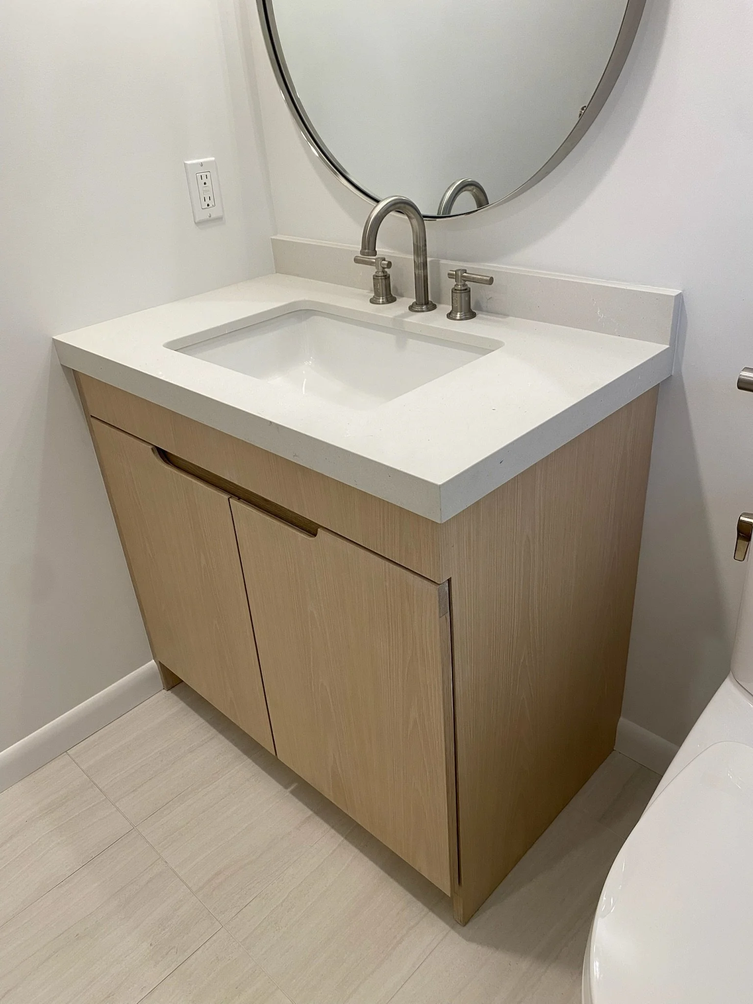 Bathroom vanity with a white countertop, a rectangular sink, a round mirror above, and wood cabinets below.