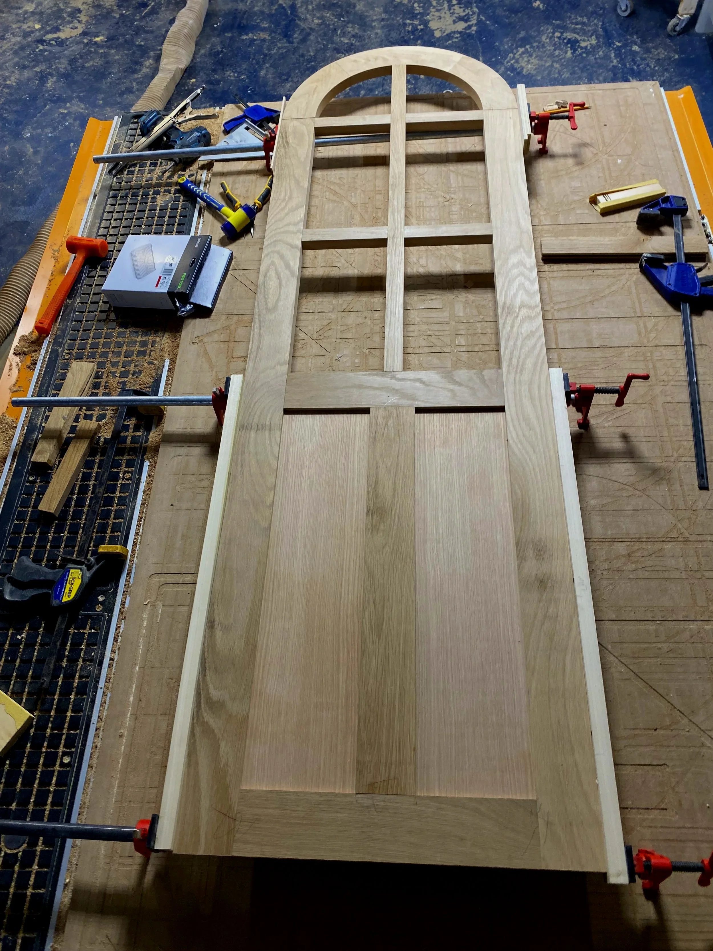 Wooden door frame being assembled on a workbench, with clamps, tools, and woodworking supplies surrounding it.