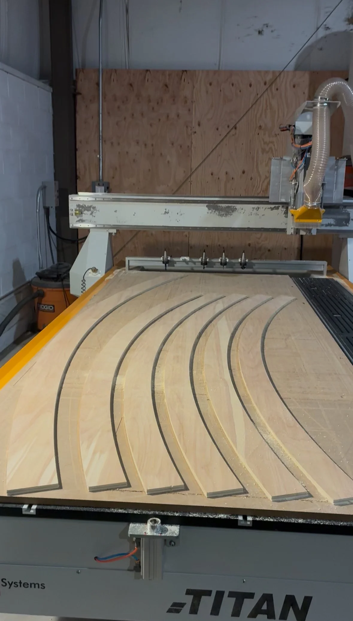 A CNC machine with curved wooden pieces on its surface, likely in a woodworking shop, with a wooden wall in the background.