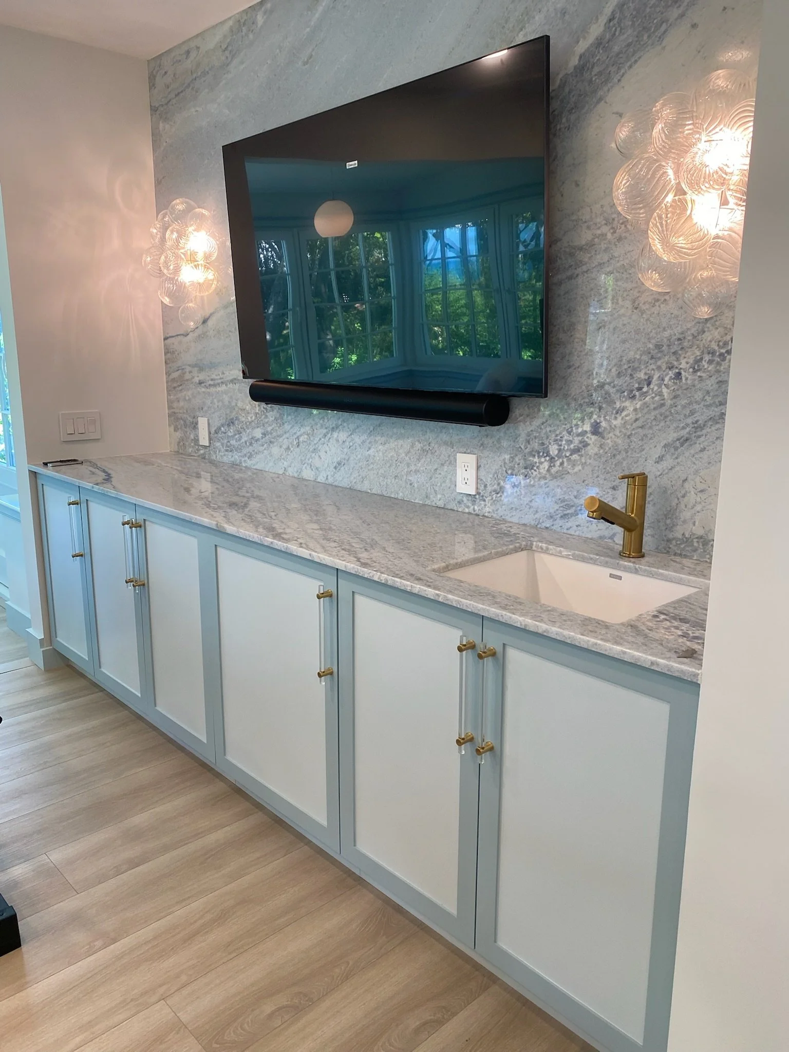A modern kitchen with light blue cabinets, a grey and white marble countertop, a small white sink with a gold faucet, a wall-mounted TV, and decorative wall lighting.