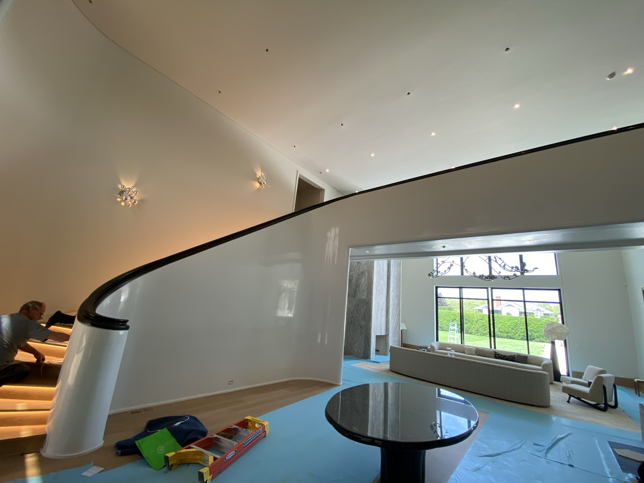 Living room with large windows, modern chandelier, white sofa, and armchairs, under construction with tools and supplies on the floor. Stairs. Millwork