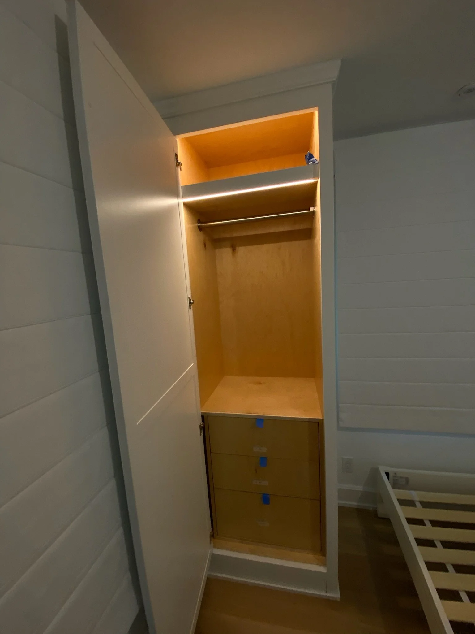 Unfinished built-in wardrobe with open doors showing an empty space, shelves, and drawers underneath, in a room with white wall paneling and hardwood flooring.