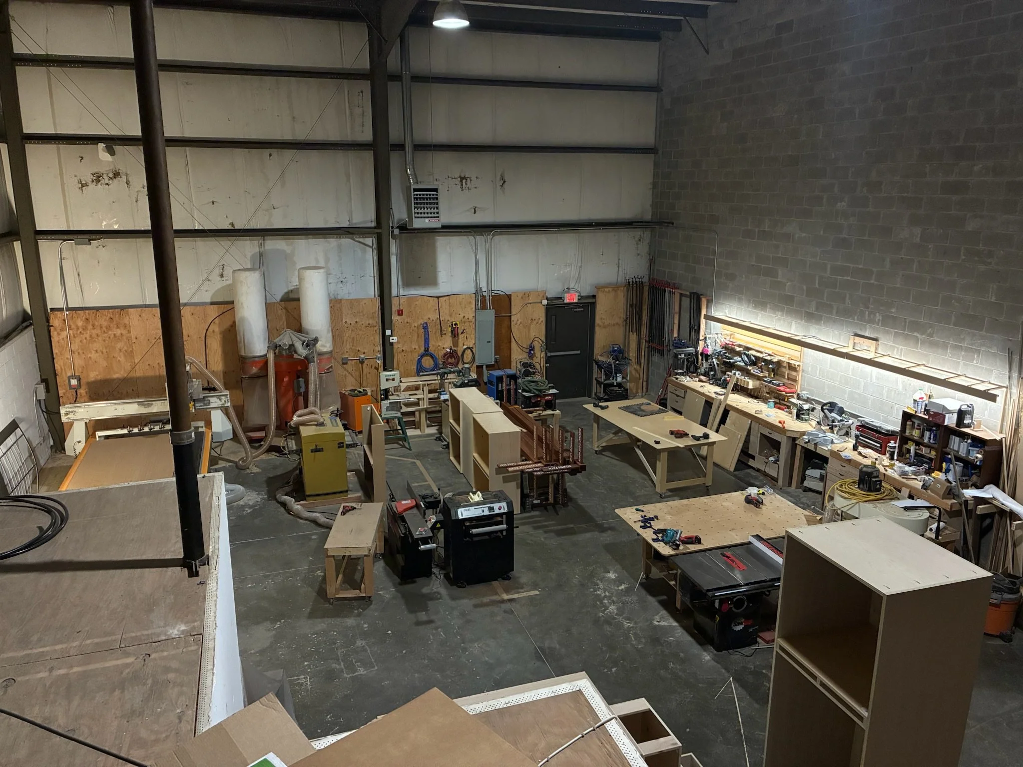 Workshop with woodworking tools, workbenches, and storage shelves in an industrial space with concrete and cinder block walls.