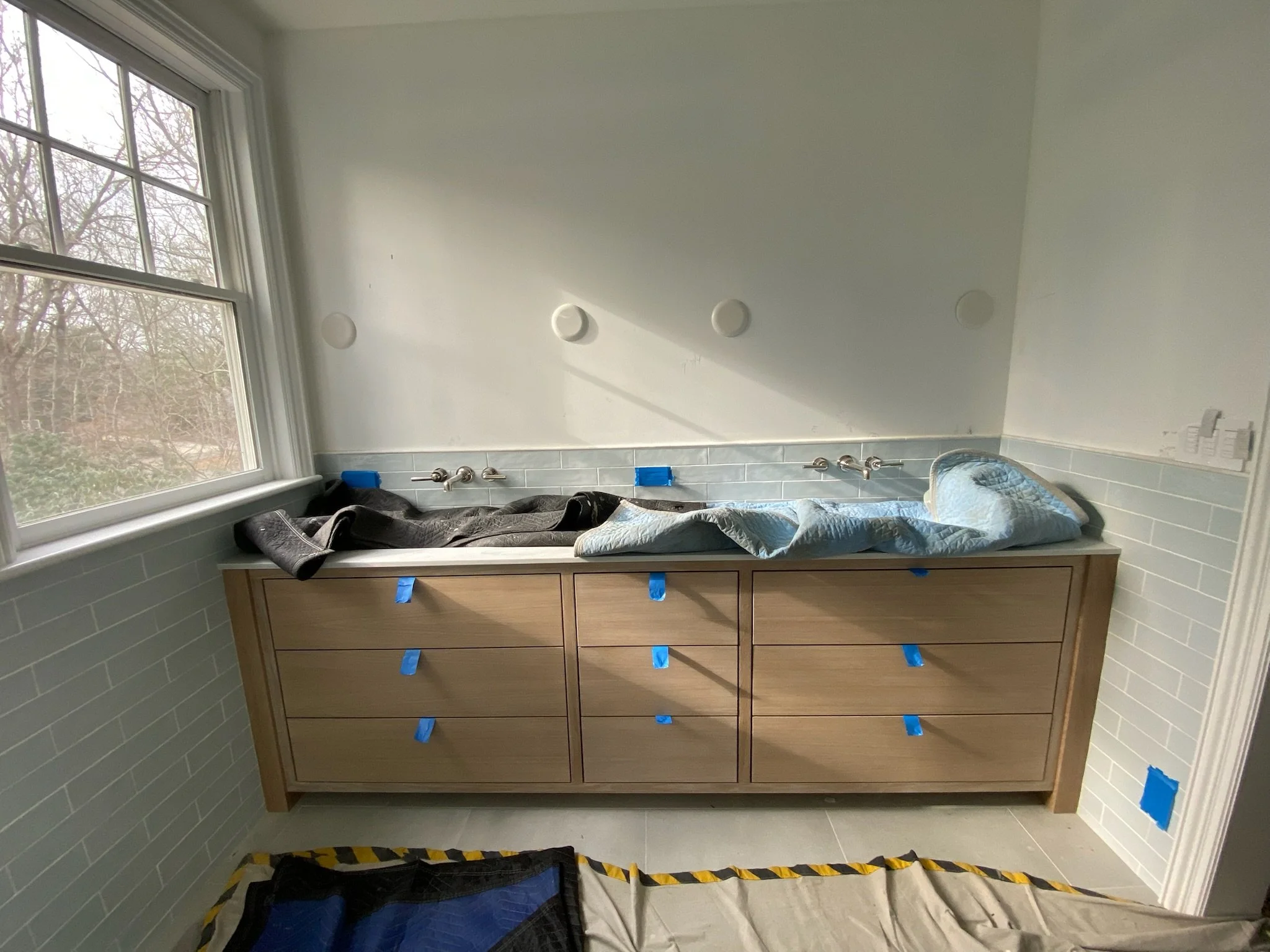 Bathroom vanity with a blue tape on drawers, a window on the left, and a countertop with items covered by cloths.