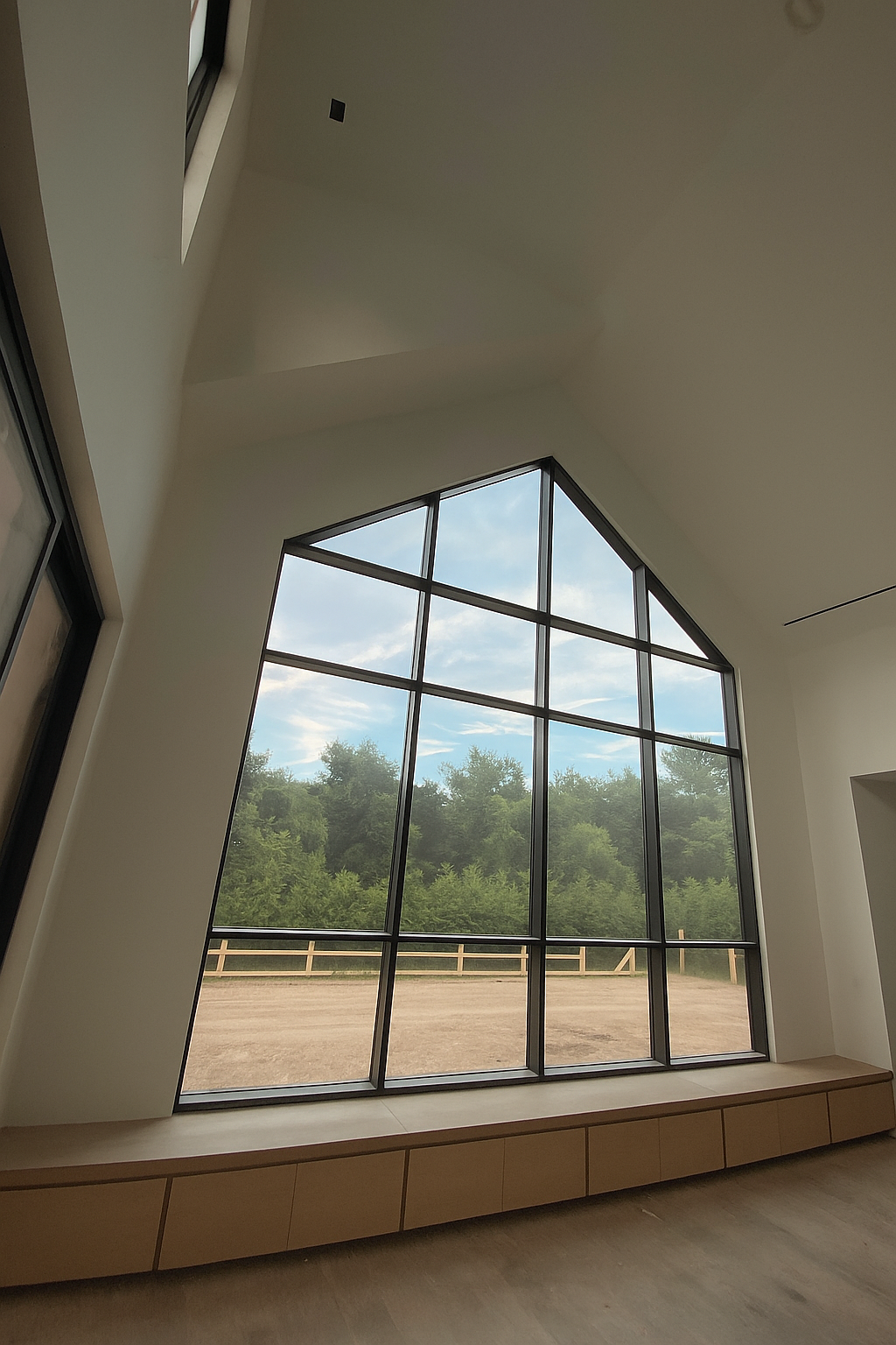 Interior view of a modern room with large, triangular window showing outdoor trees and fence, light-colored walls, and a built-in wooden bench beneath the window. cabinet renovation. woodwork.