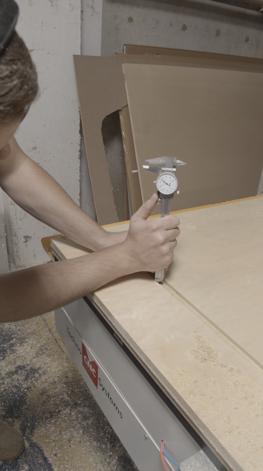 A person using a dial caliper to measure a piece of wood in a woodworking shop. Several sheets of plywood are stacked in the background, and the measuring device is held vertically on the wood surface.