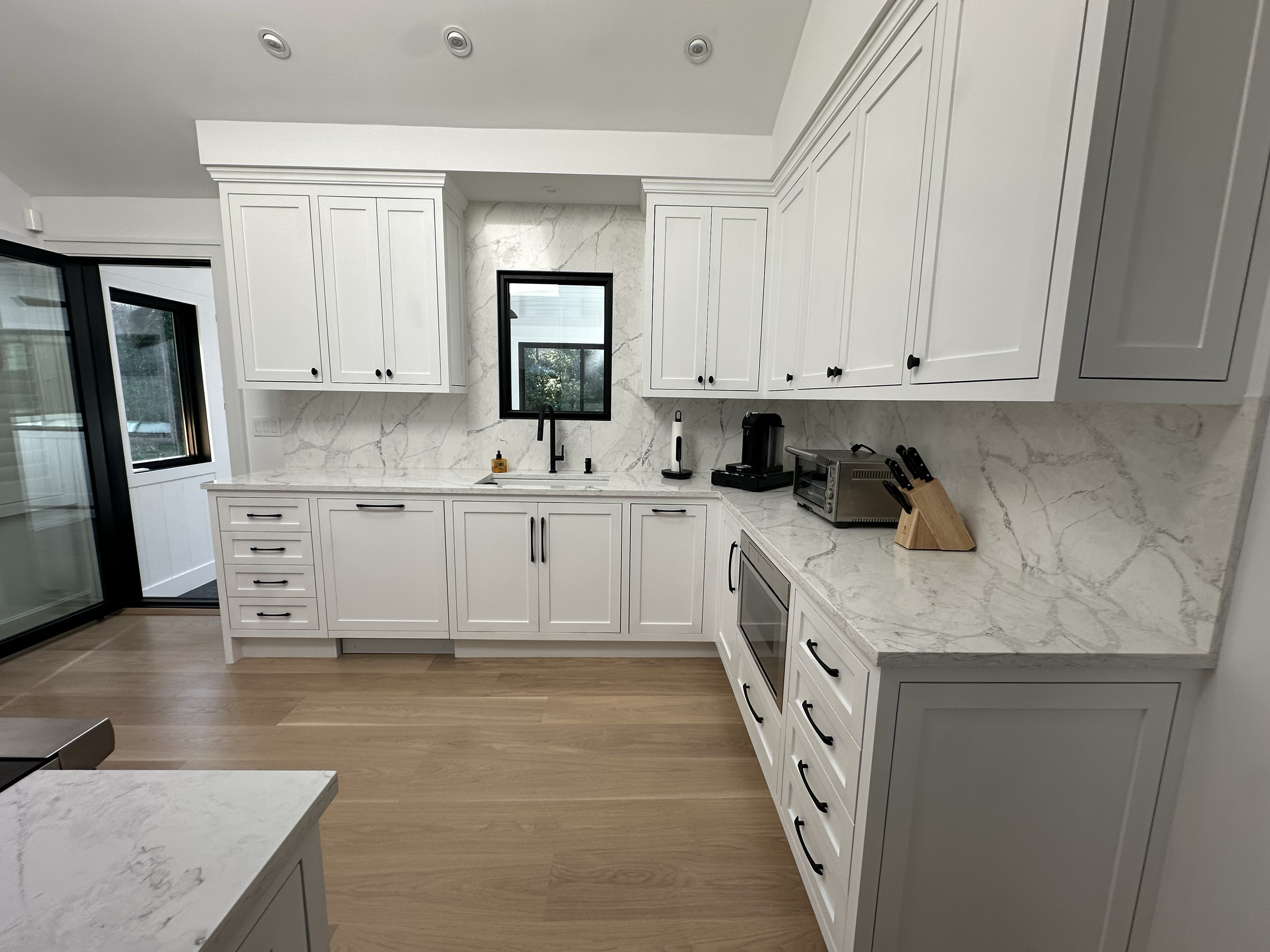 Modern kitchen with white cabinets, marble countertops, black hardware, a small window above the sink, and kitchen appliances on the counter.
