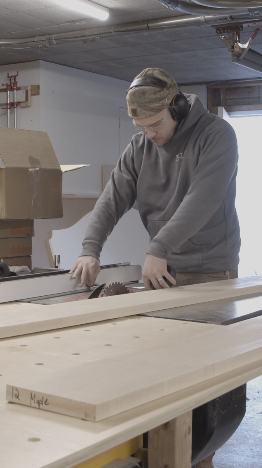 A man wearing safety goggles, headphones, a hooded sweatshirt, and a camouflage cap cutting wood with a table saw in a woodworking shop.