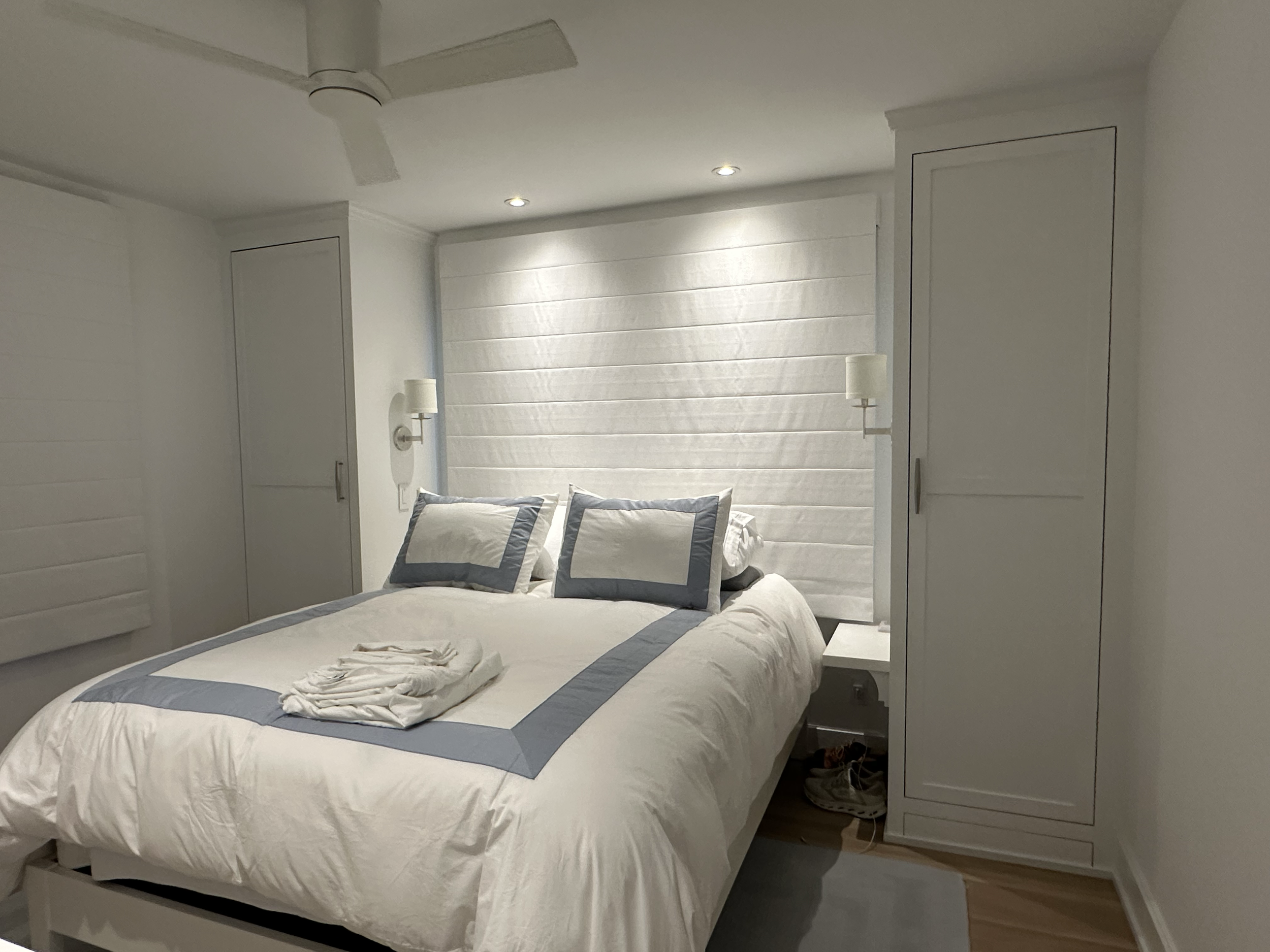 A neatly made bed with white and blue bedding, two pillows, and a folded towel in a bright, minimalist bedroom with white walls, built-in closets, and wall-mounted lamps.