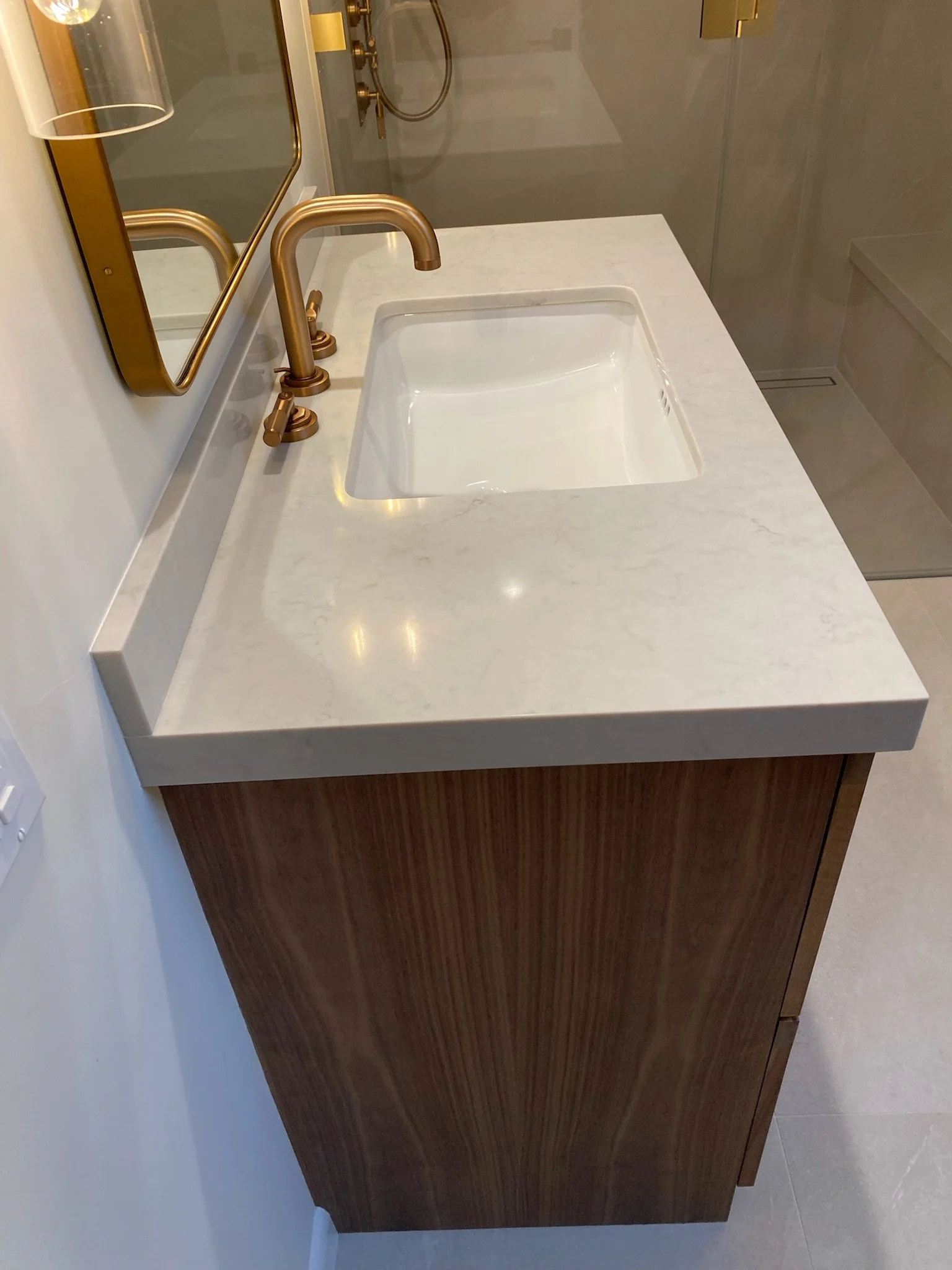 Bathroom vanity with a white marble countertop, a built-in white sink, and a gold faucet, with a mirror and a shower area visible in the background.