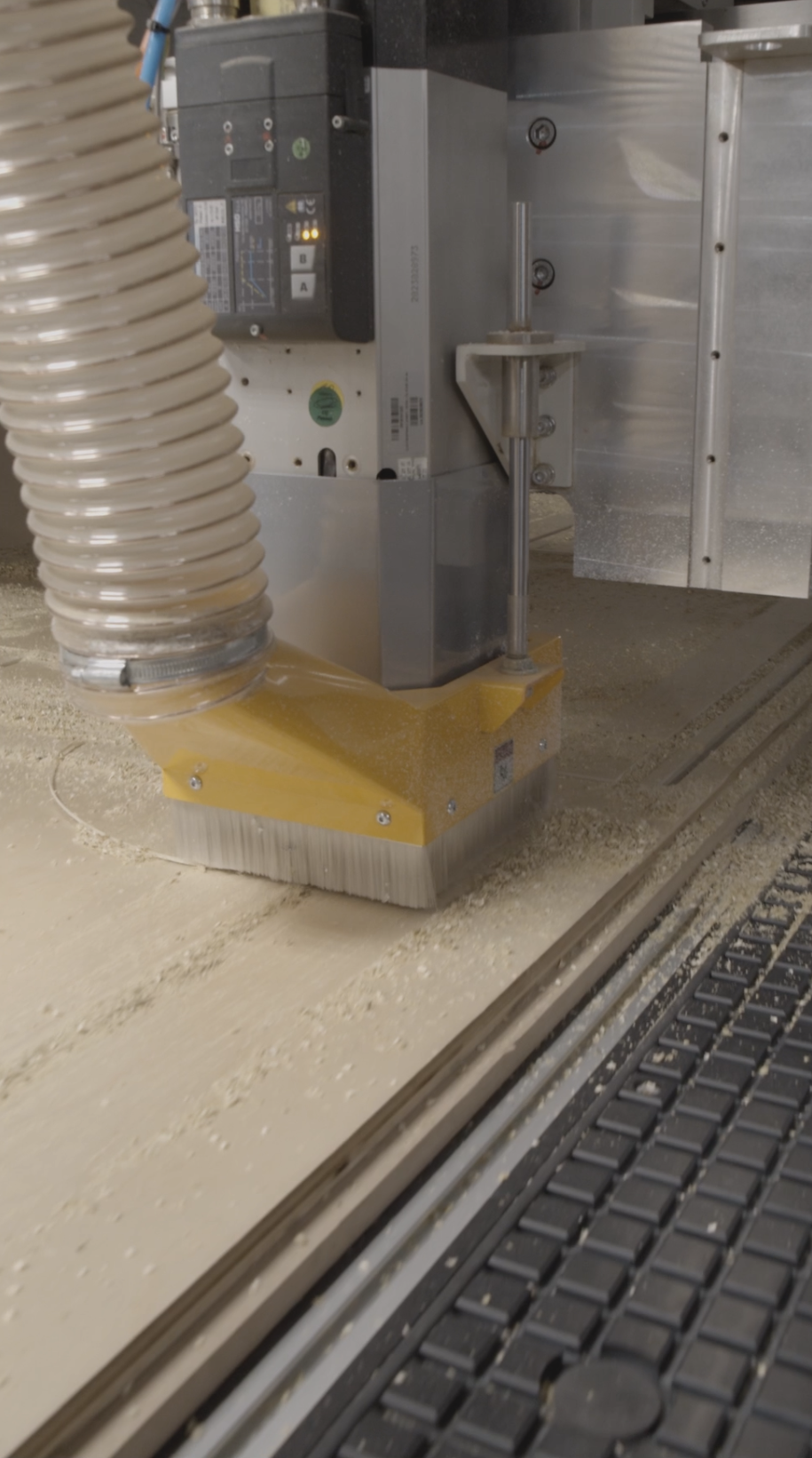 Close-up of woodworking CNC machine cutting a wooden surface with dust and shavings around, with the machine's dust collection hose attached.