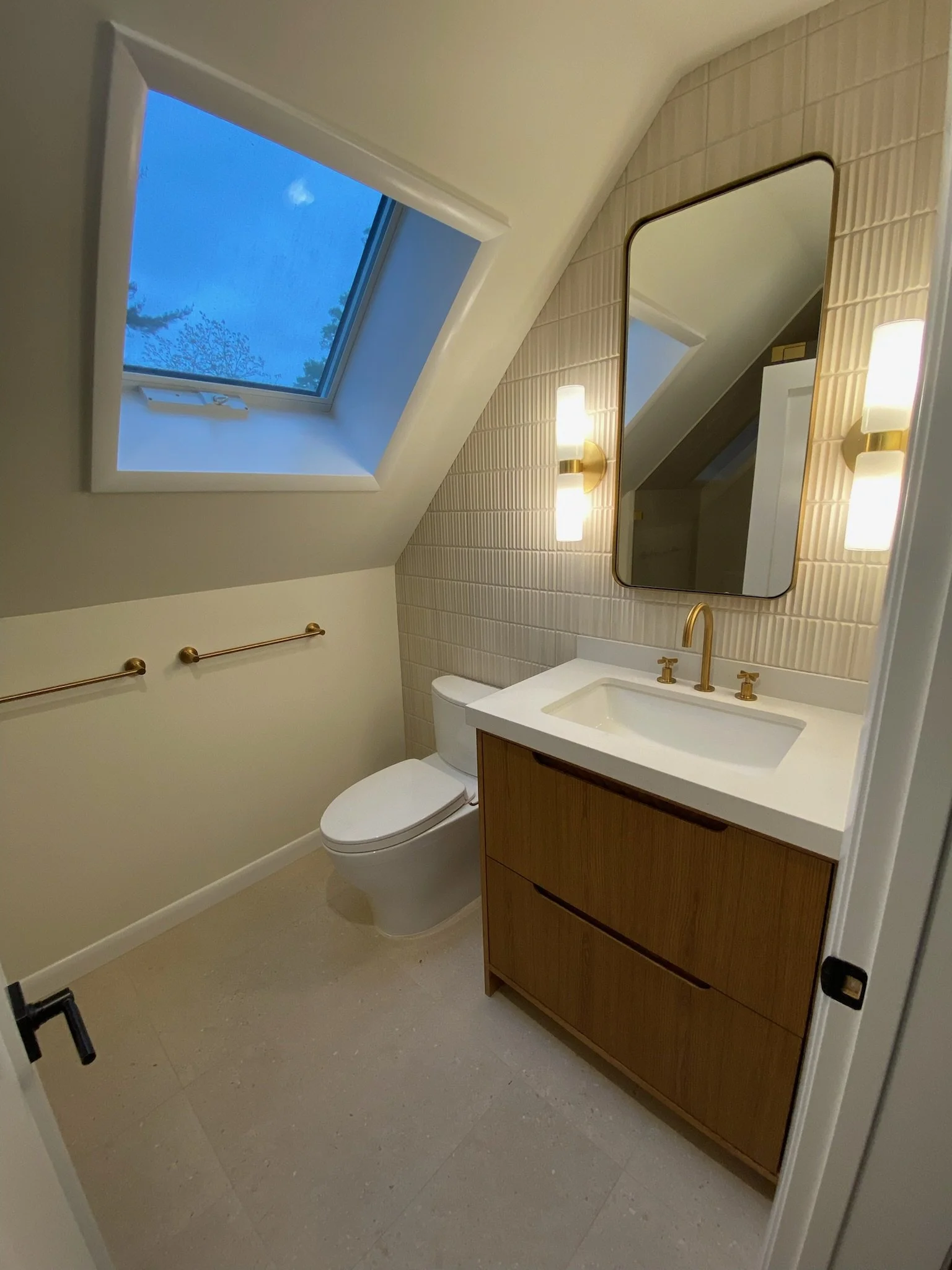 Small bathroom with a sloped ceiling, skylight window showing a blue sky, a modern toilet, a wooden vanity with a white sink, gold fixtures, and a large mirror with gold-framed wall sconces on each side.