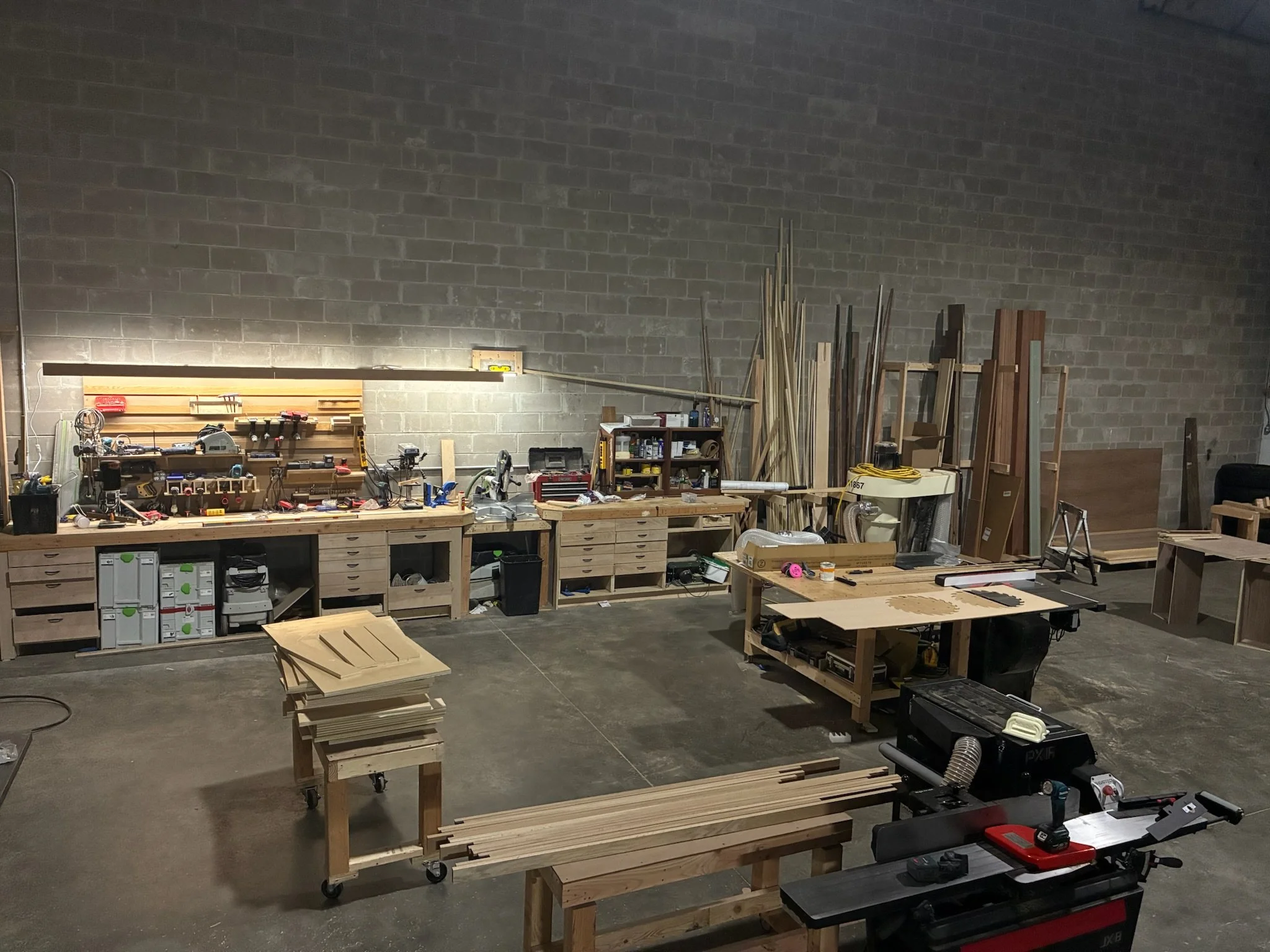 A woodworking workshop with various tools and wooden materials, including workbenches, shelves, and stacks of wood in a spacious, industrial-style room.