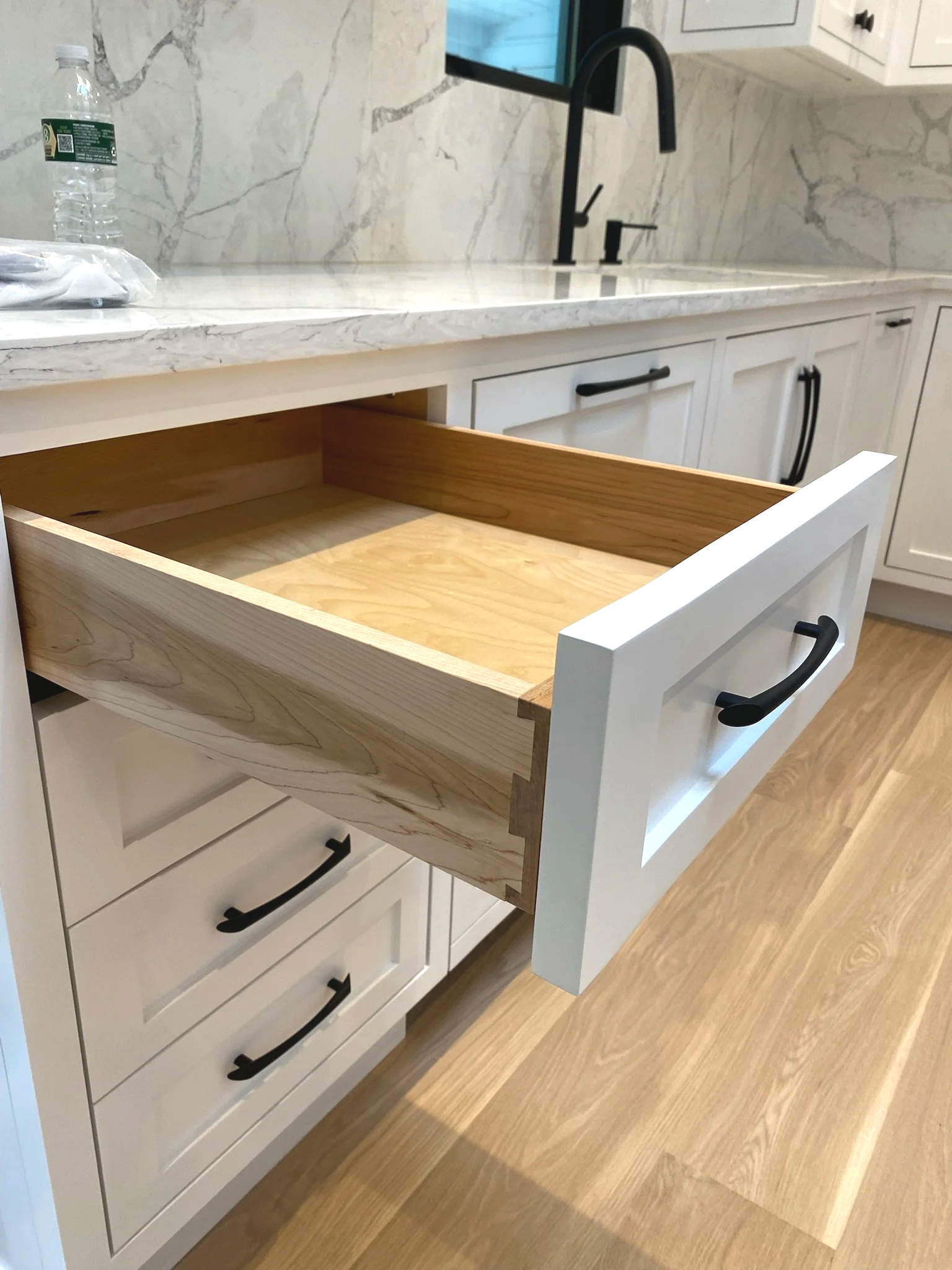 A kitchen drawer partially pulled out from white cabinetry, showing the unfinished wood interior. The kitchen has a marble countertop and backsplash, a black faucet, and black handles on other drawers and cabinets. A water bottle is placed on the countertop.