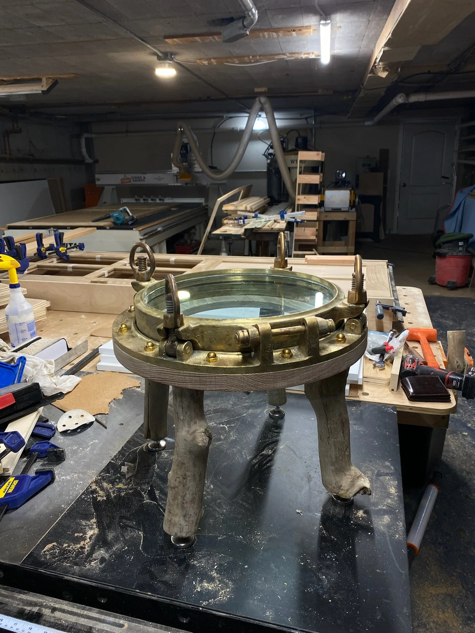 A workbench in a woodworking shop with a brass and glass porthole window mounted on a wooden stand. There are various tools and materials scattered around, including clamps, a ruler, a drill, and spray bottles.  Wooden. table.