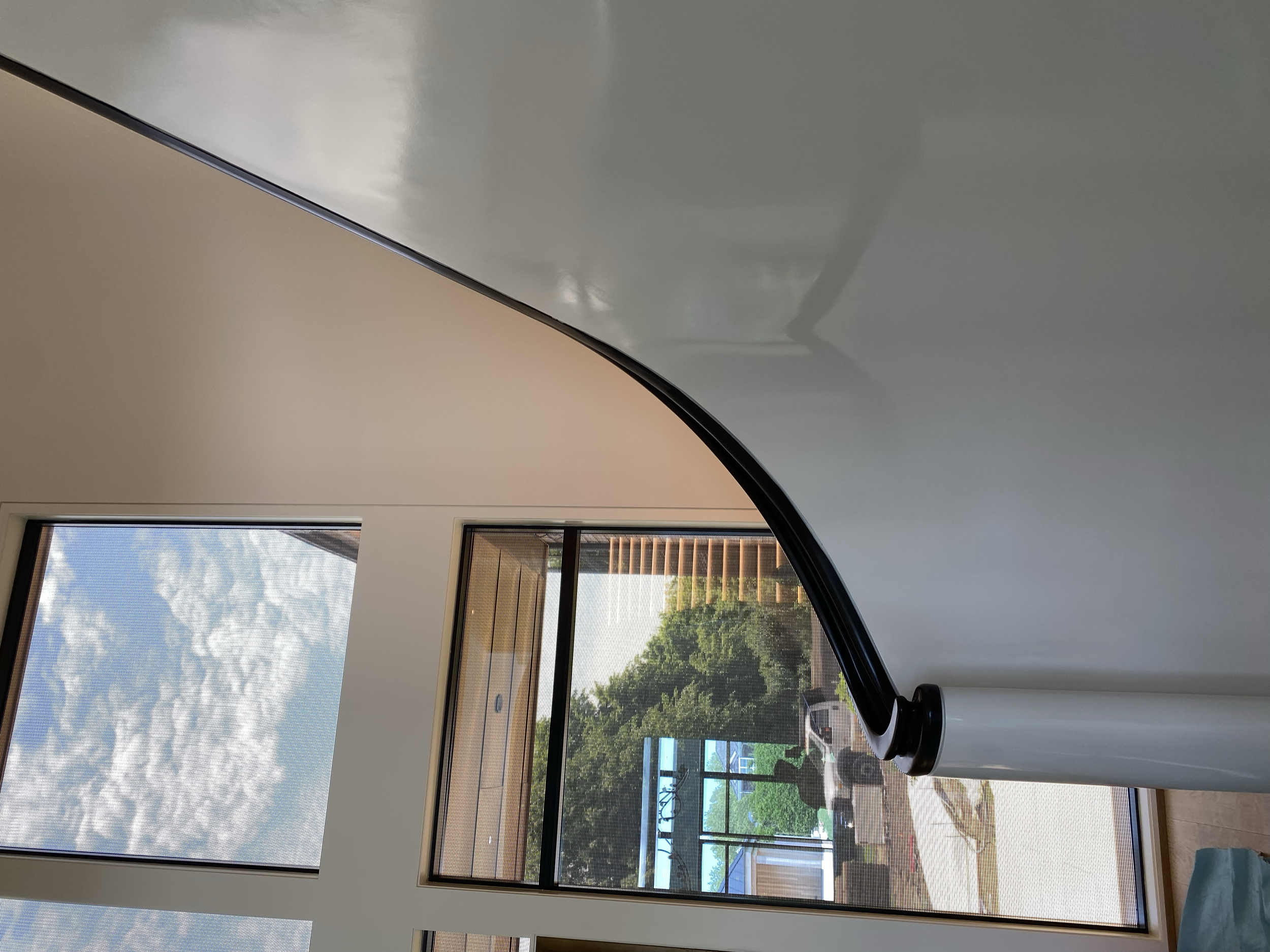Interior of a room with large windows showing an outdoor view of trees, a building, and a blue sky with clouds. A yoga or massage table with a rolled-up sheet or towel on it is in the foreground.