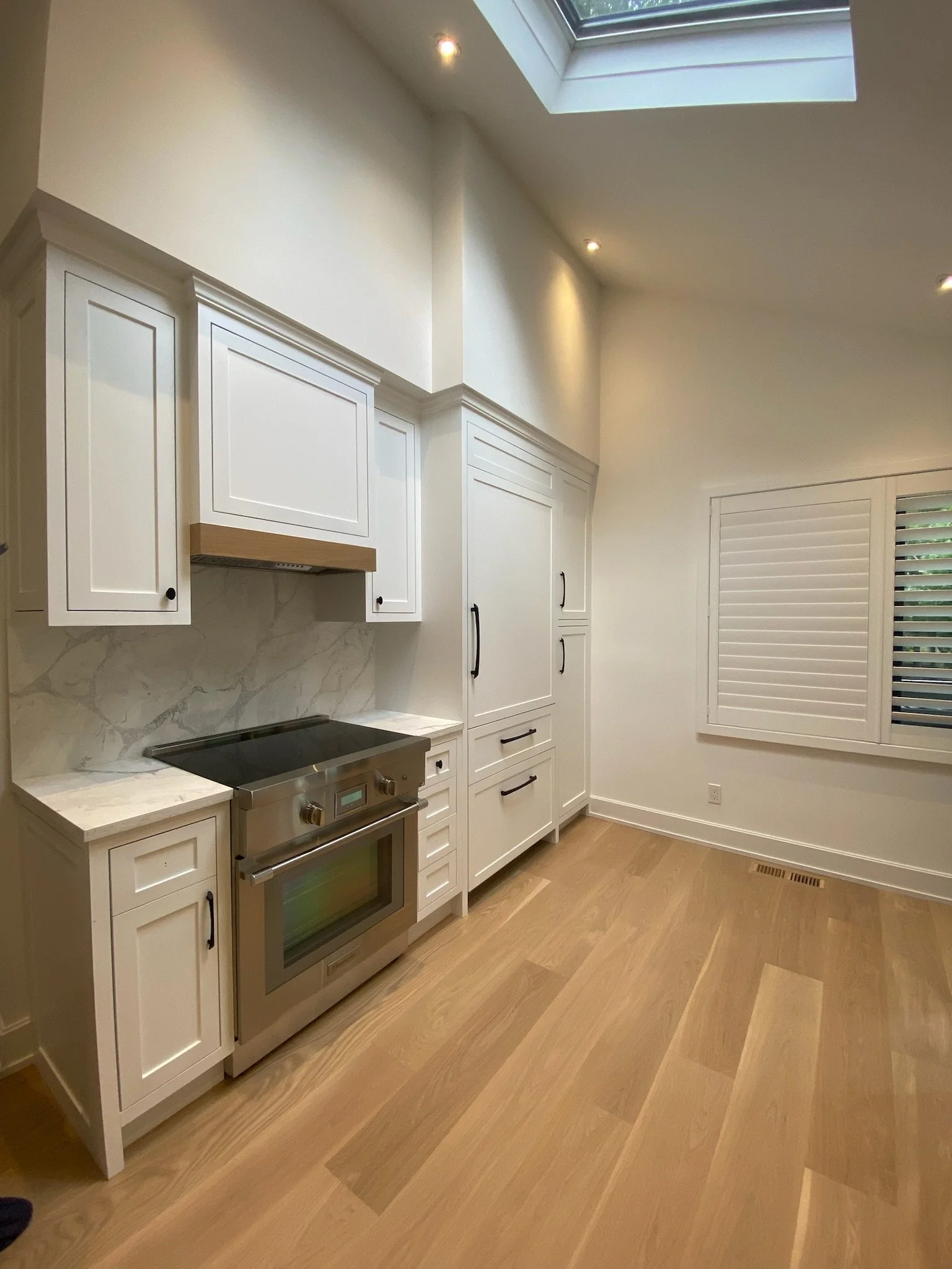 Empty modern kitchen with white cabinets, a marble backsplash, a stainless steel oven, wooden floors, a large window with white shutters, and a skylight.