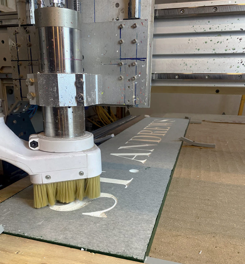 Close-up of a CNC machine engraving white text on a gray surface, with a yellow brush cleaning the surface.