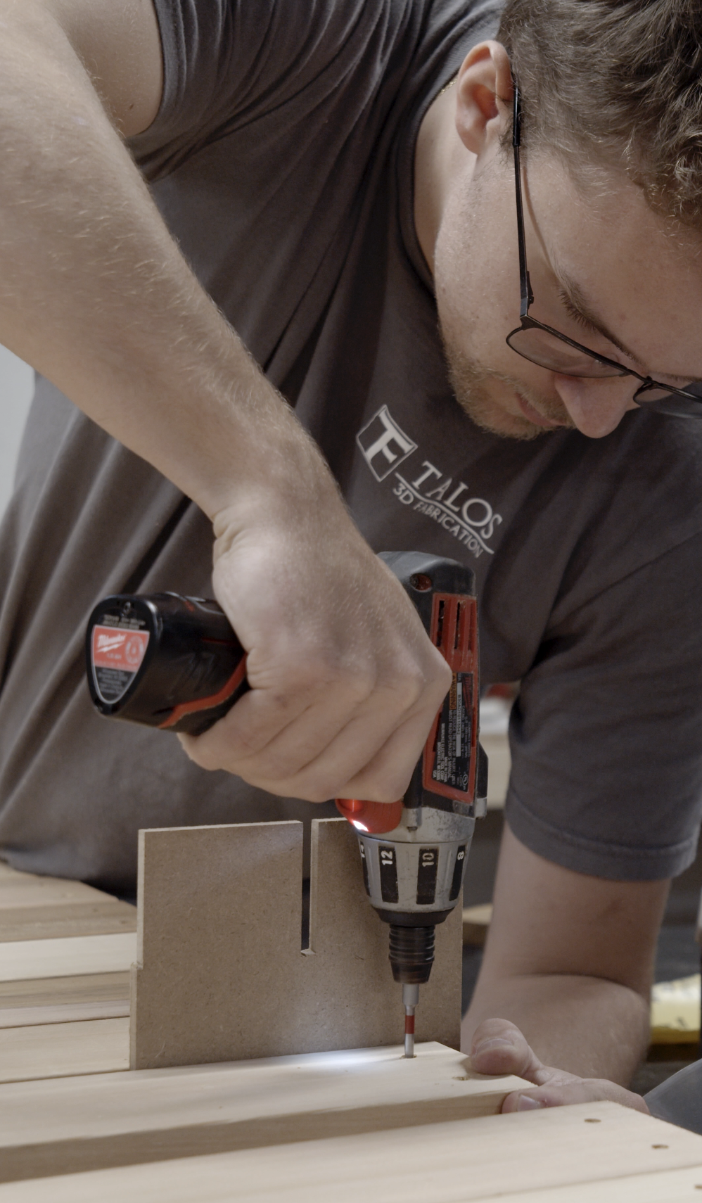 A man wearing glasses and a gray T-shirt is using a cordless drill to drill into a piece of wood in a woodworking shop. Cabinet.