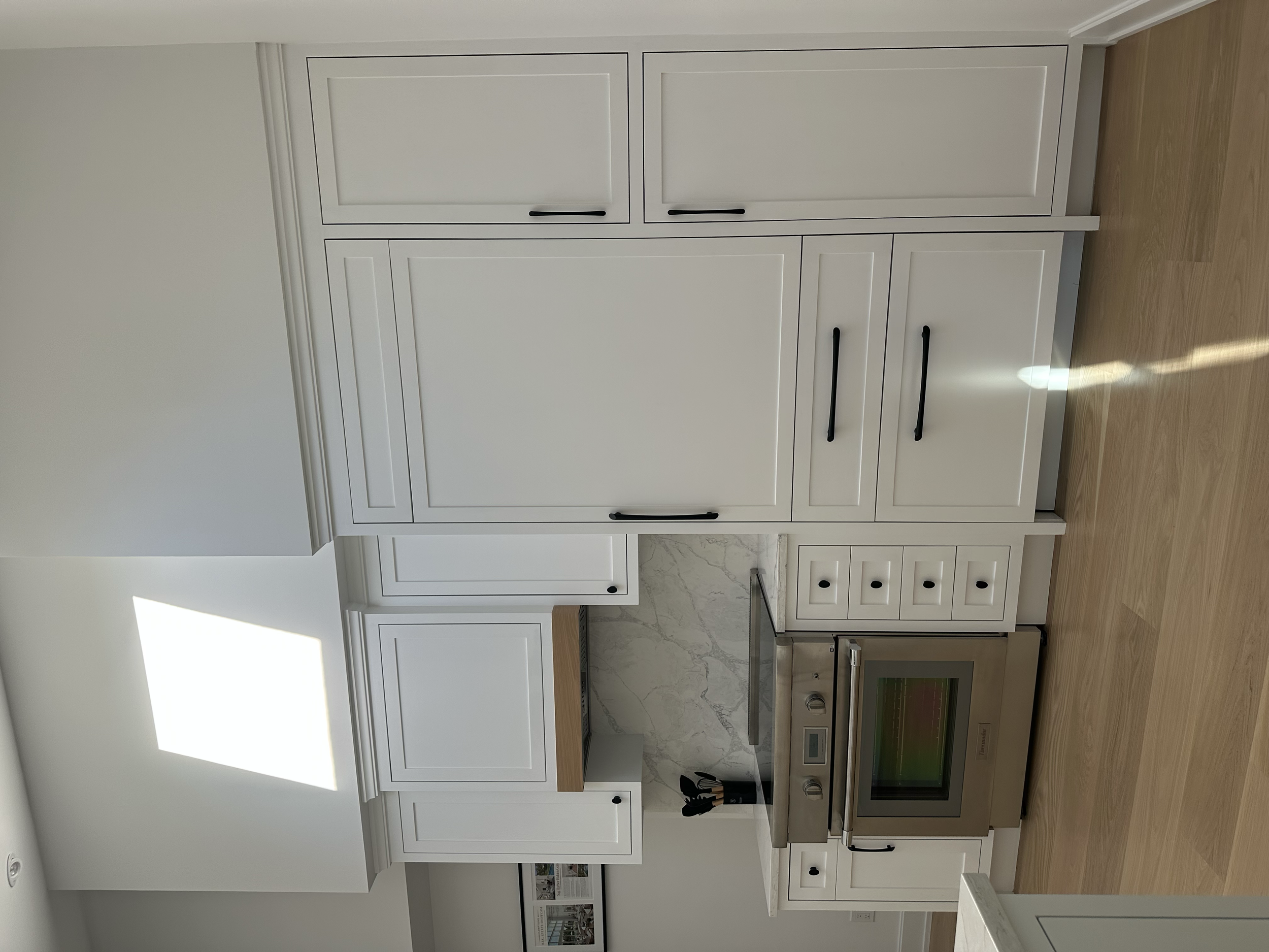 White kitchen cabinets with black handles, a marble backsplash, an oven, and hardwood flooring.