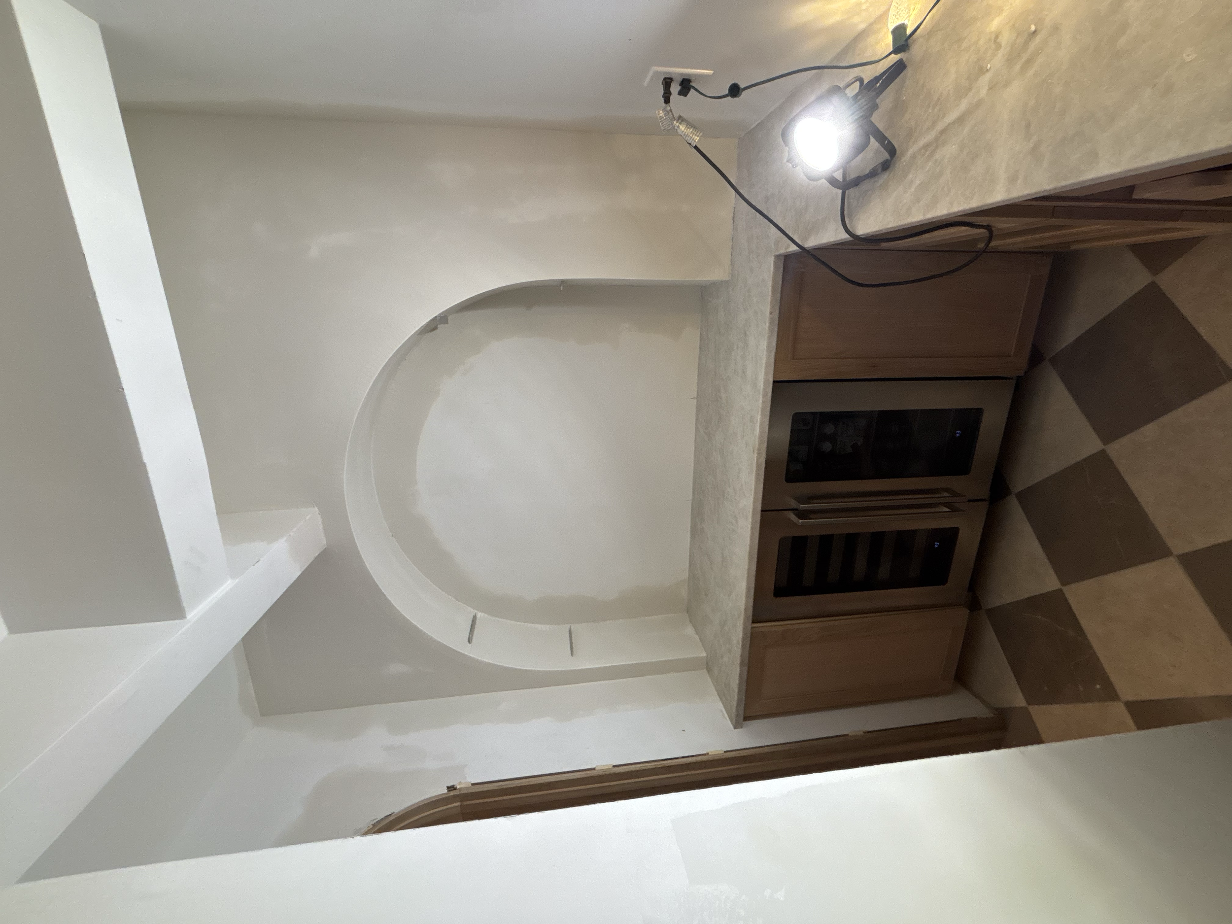 Kitchen with beige granite countertops, wooden cabinets, and a wine cooler. A work light is plugged in and placed on the counter, illuminating the space. The floor has a brown and tan checkerboard pattern. The wall has an arched niche and appears to 