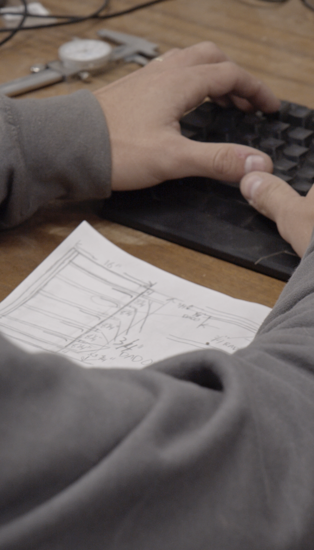 A person working on a sketch or blueprint at a wooden table, with a caliper and other tools nearby.