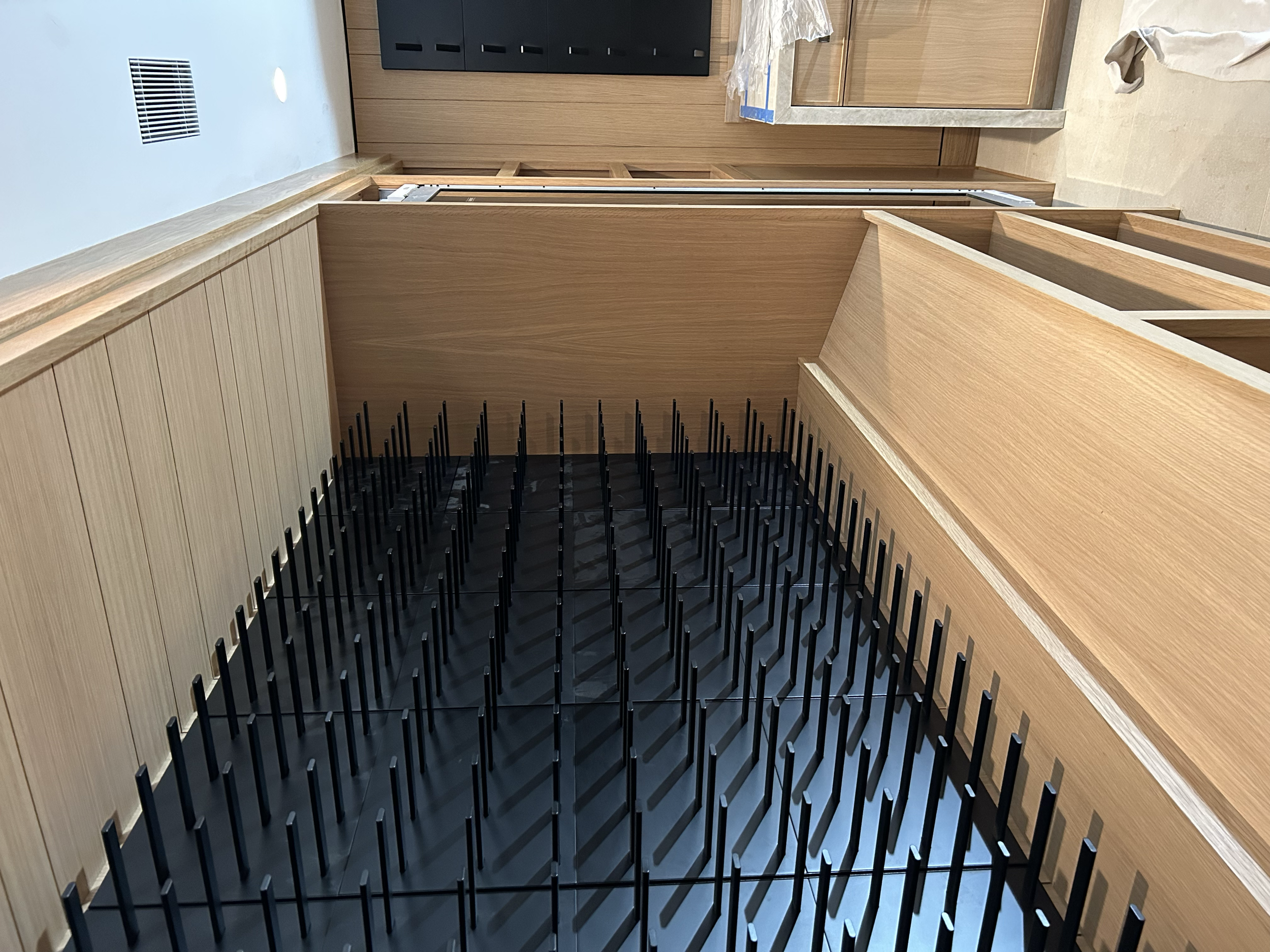 Interior of a kitchen under construction with wooden cabinets, a countertop, and black metal wine racks in the foreground.