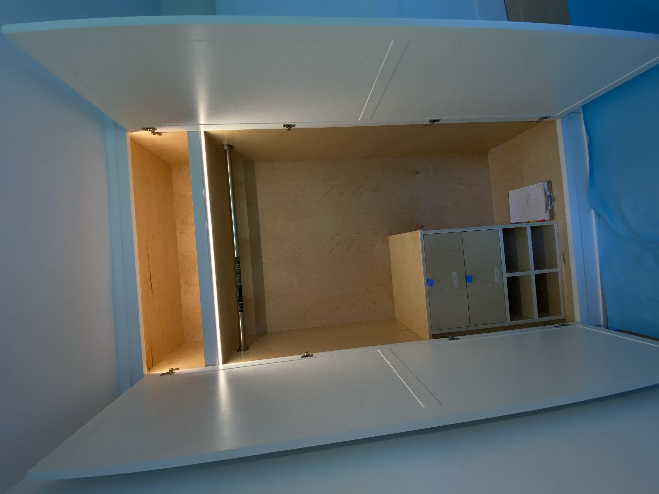 Interior of a small, unfinished closet with wooden shelves and open cabinet space, with a clipboard on the floor, and a blue plastic covering on the right side. Storage.