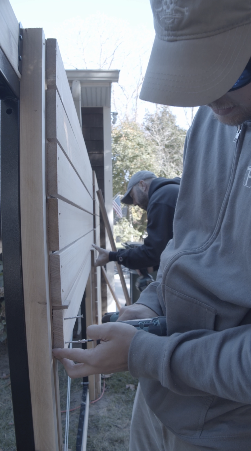 Two men working on a woodworking project outdoors, attaching horizontal wooden slats to a vertical structure with screws. Fence.