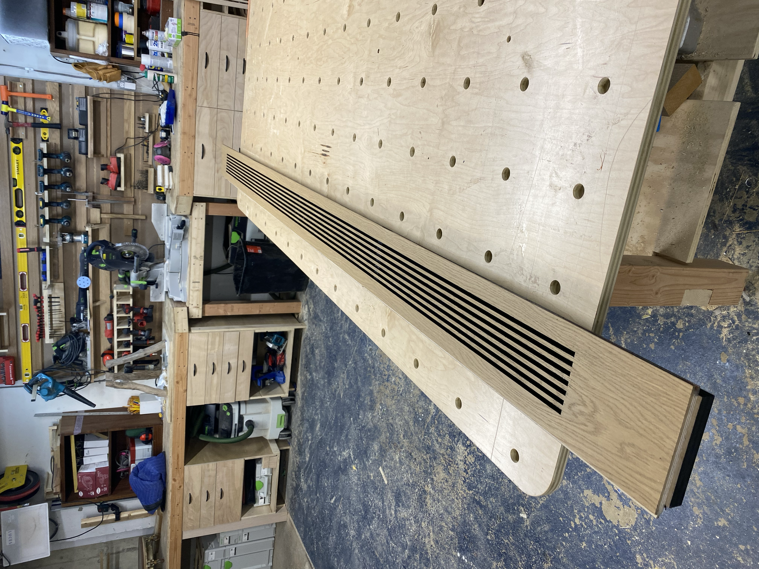 Woodworking workshop with a large workbench, various tools, and a long curved piece of wood with black slots.