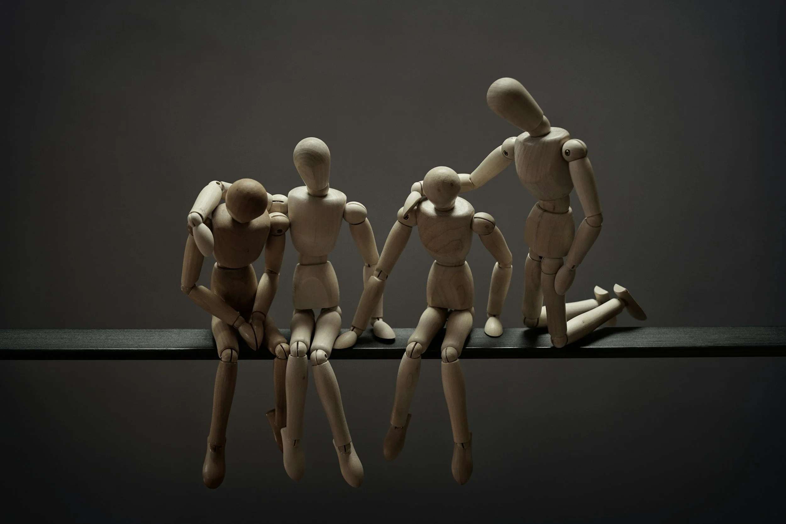 A group of wooden artist mannequins are posed on a reflective surface against a dark background, with some mannequins appearing to bow, kneel, or lean.