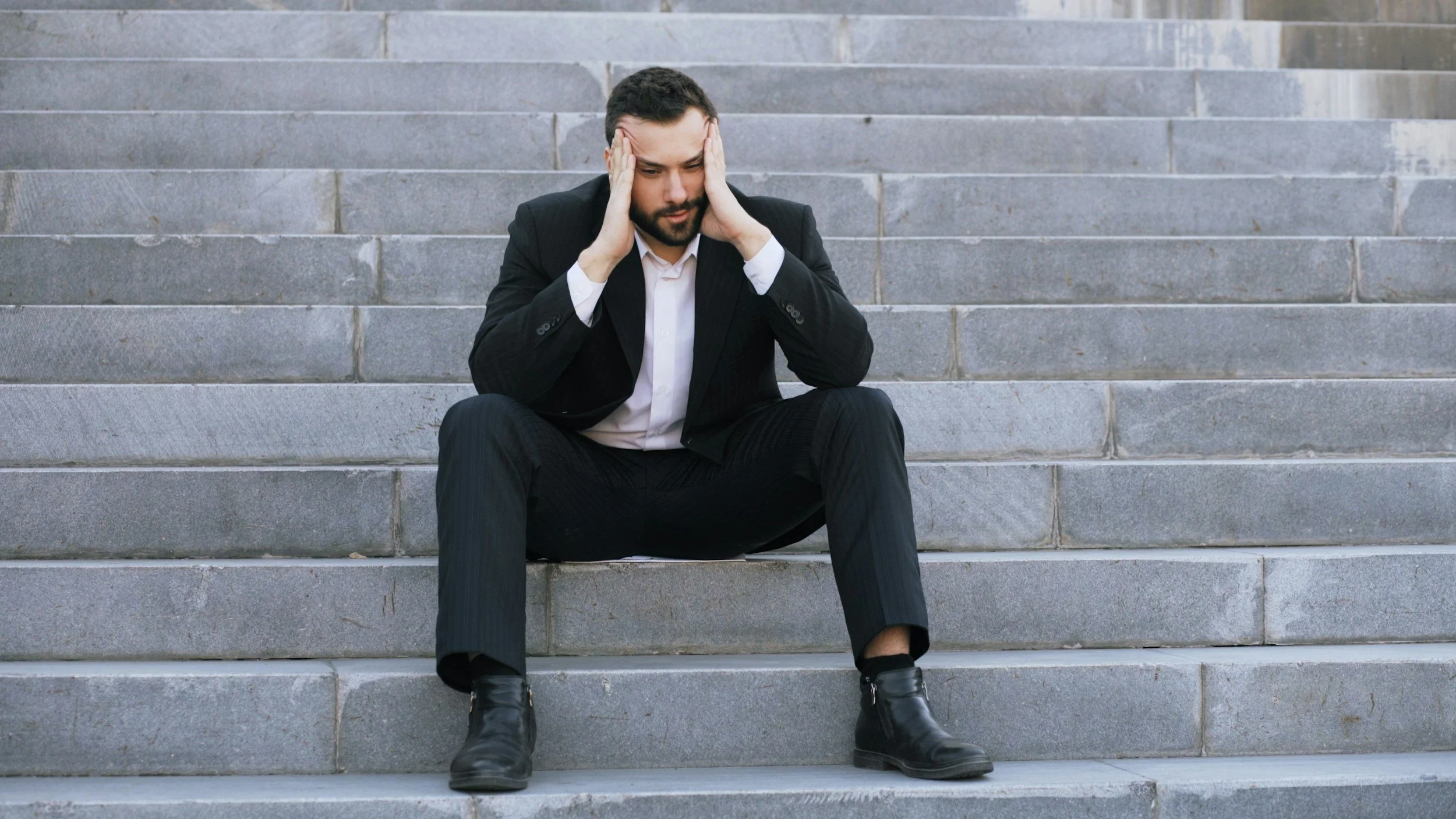 A man wearing a business suit sitting on grey stone steps, holding his head with both hands, appearing stressed or overwhelmed needing therapy for performance anxiety.