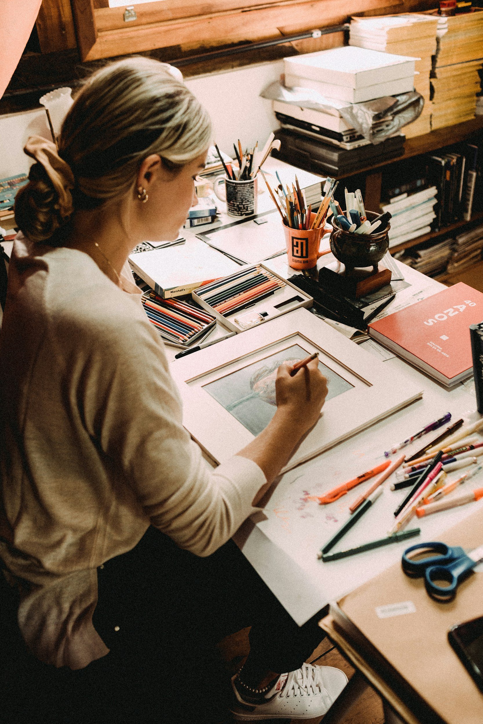 A person with blonde hair tied back, wearing earrings, sketching in a sketchbook at a cluttered table with markers, scissors, and colored pencils, in a cozy, wood-paneled room.