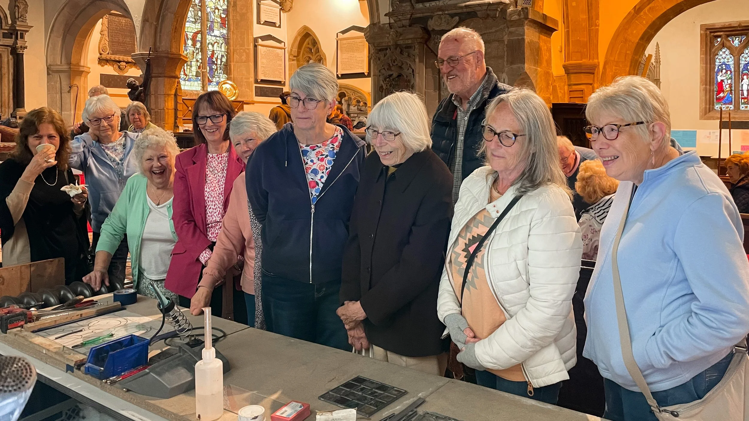 A group of around 12 visitors enjoying a stained glass workshop - tools in the foreground on a table