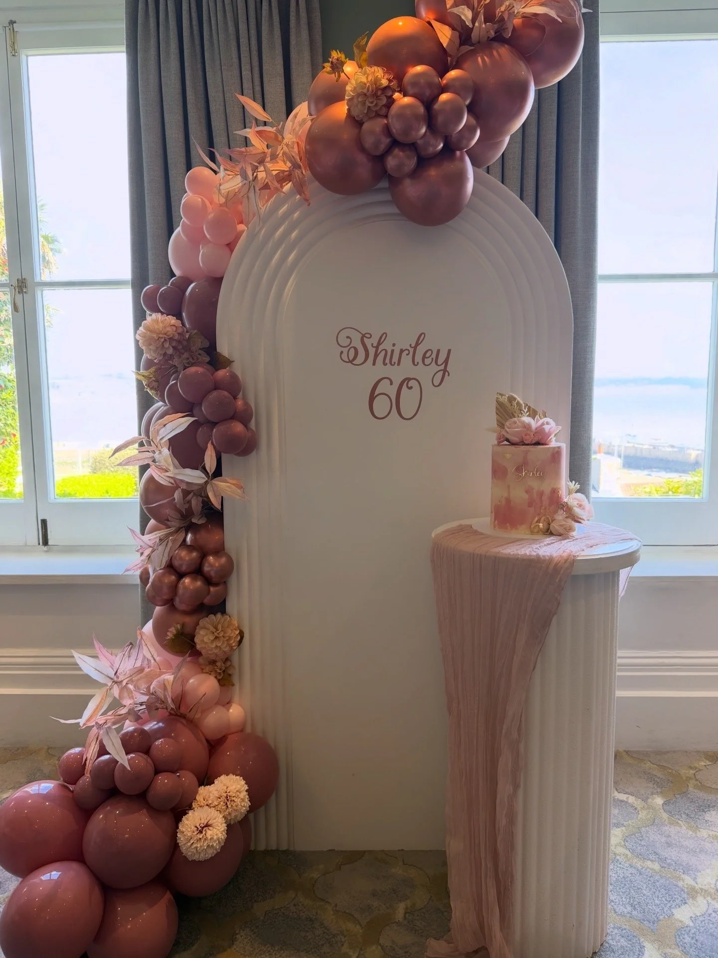Balloon decoration with clouds in white, gold, and green, a white pedestal with a cake, a religious cross on the wall, and curtains in the background.