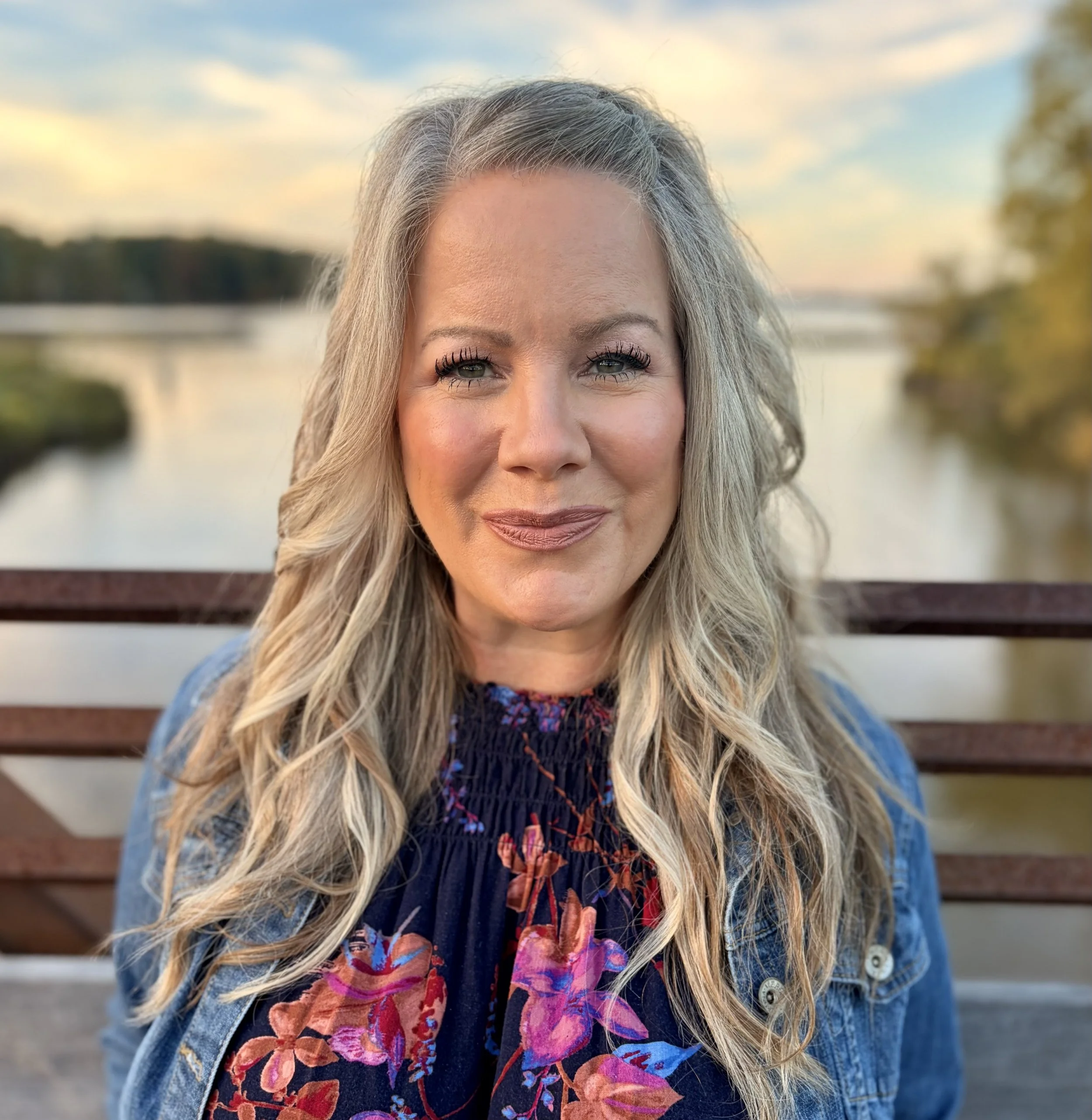 Close-up of a blonde woman smiling outdoors during sunset, with a lake and trees in the background, wearing a denim jacket and a colorful scarf.