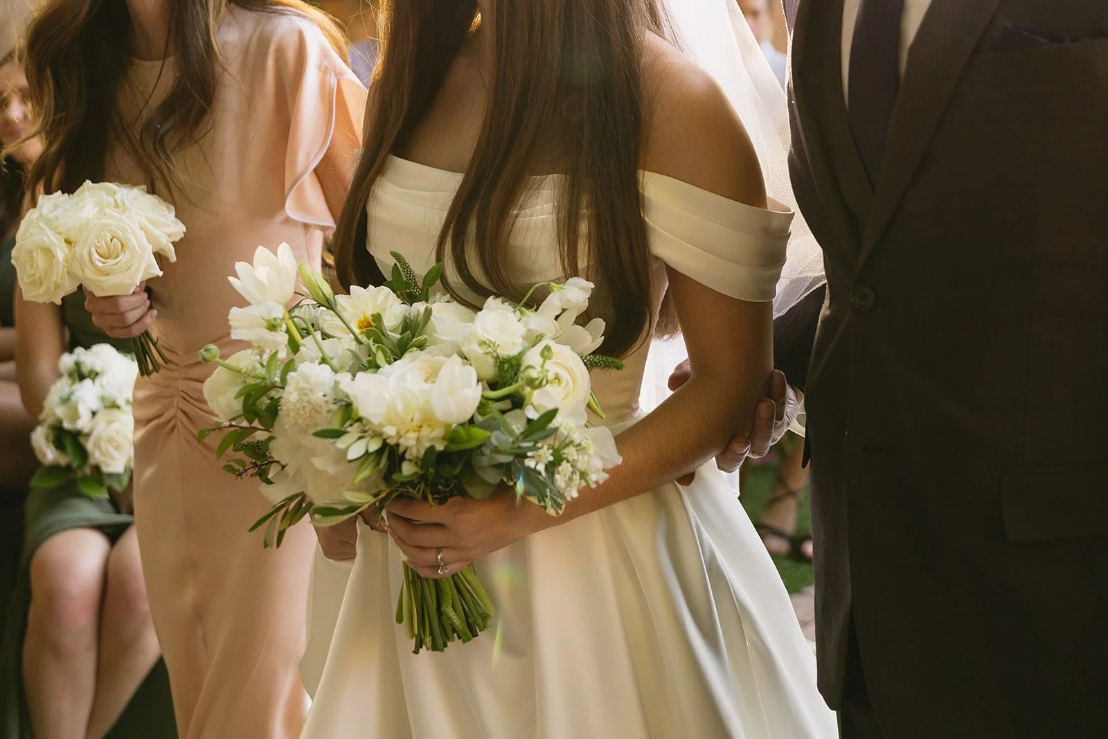 A bride holding a bouquet of white flowers at her wedding ceremony, with bridesmaids and guests in the background.