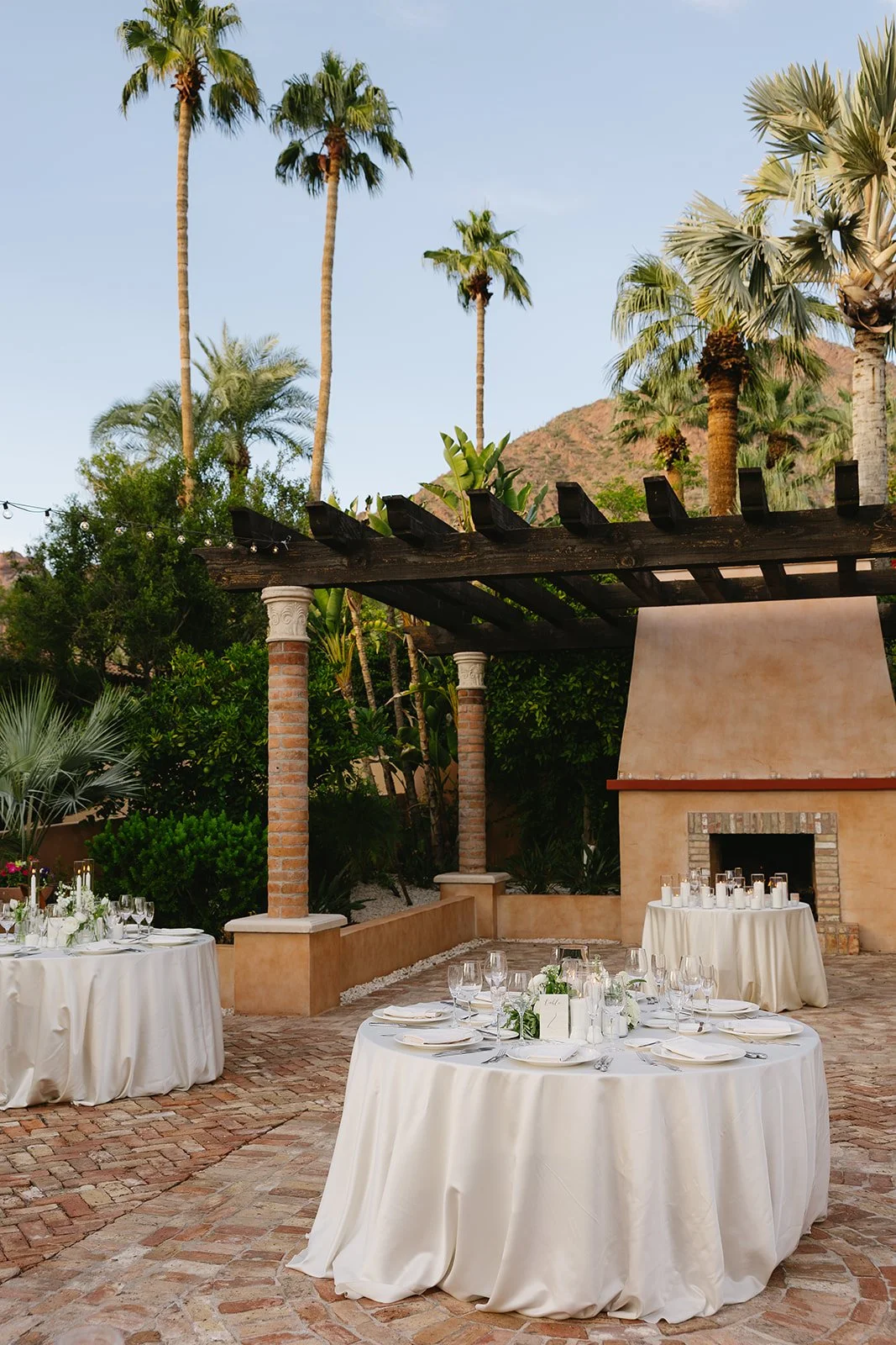 Outdoor wedding setup with round tables covered in white tablecloths, decorated with glassware, plates, and green accents. Stone fireplace and lush palm trees surround the area, with a clear sky overhead.