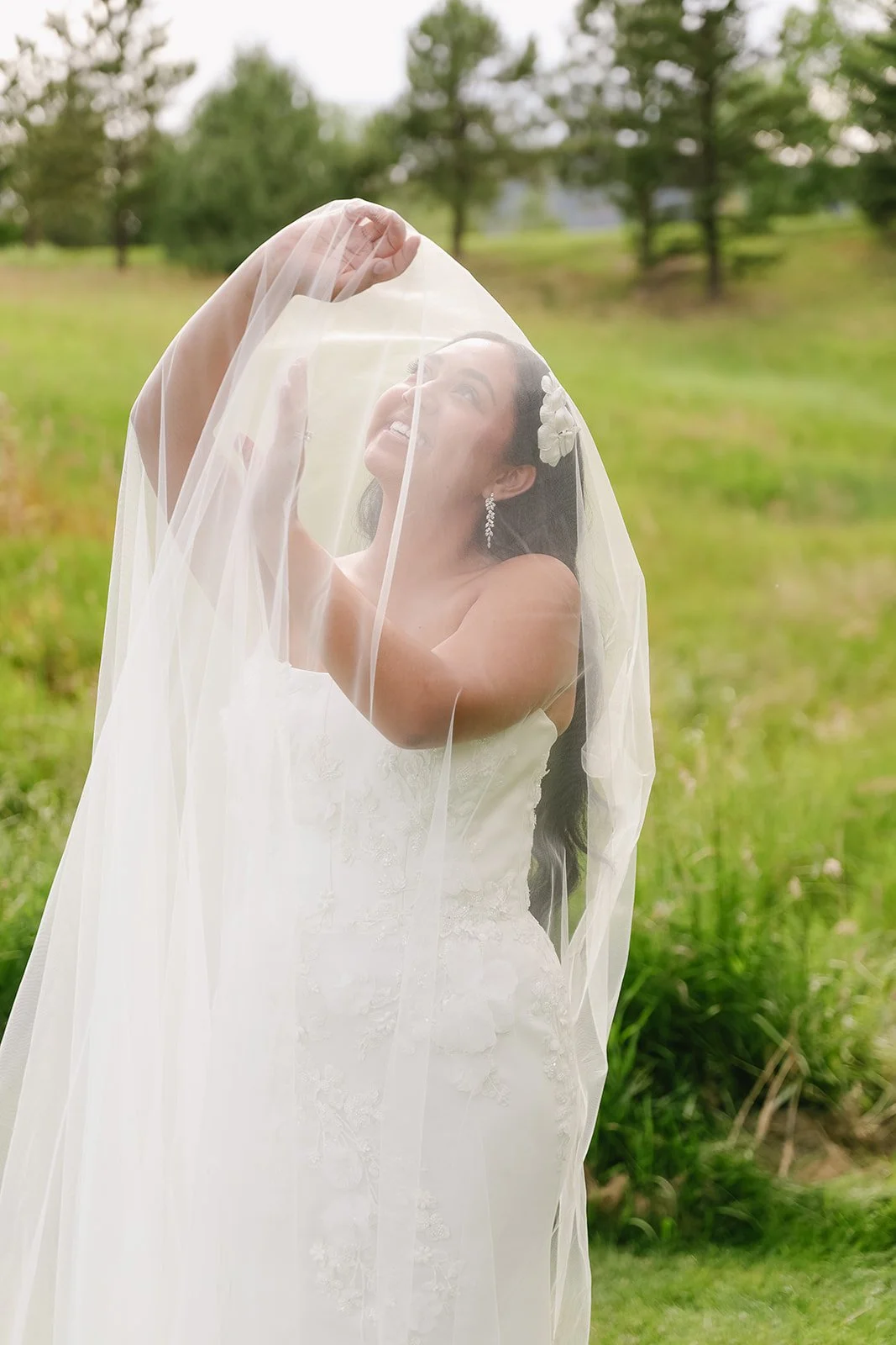 A bride smiling underneath a wedding veil outdoors with green grass and trees in the background.