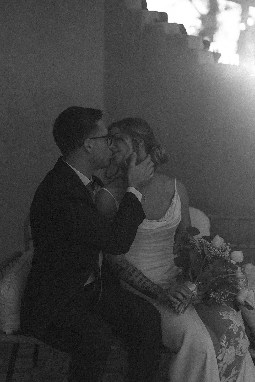 A couple sitting close together, about to kiss, in a black and white photo. The man is wearing a suit and glasses, and the woman is holding a bouquet of flowers, with tattoos on her arm, wearing a dress with spaghetti straps.