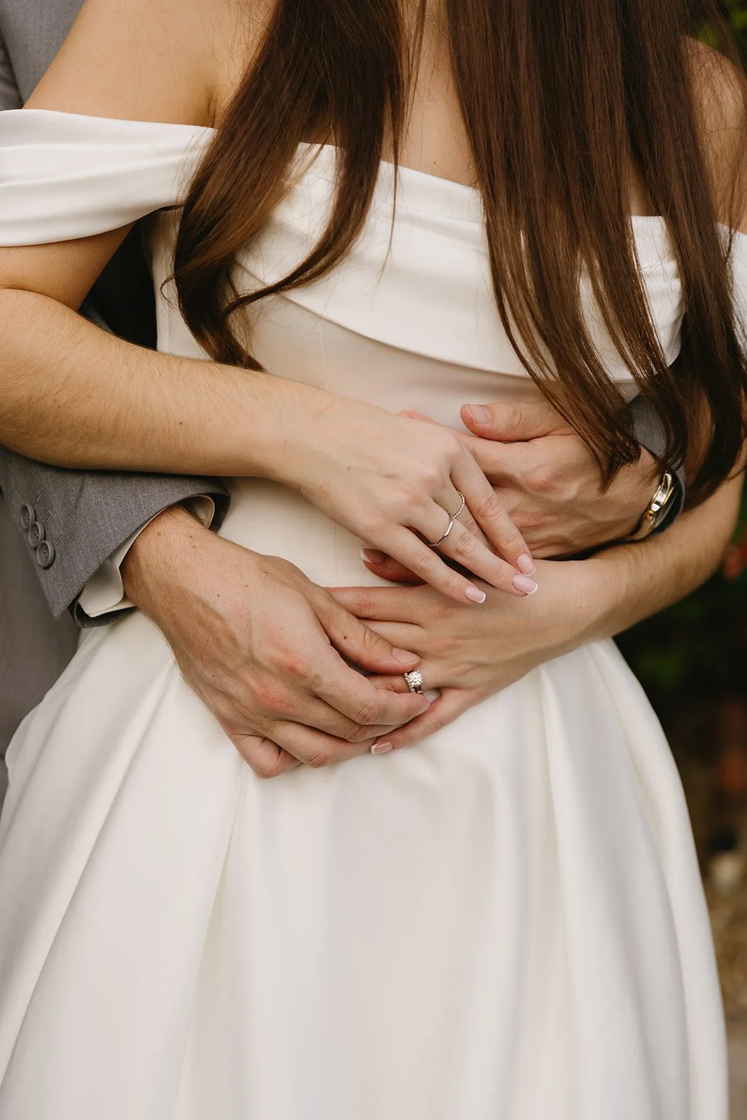 Close-up of a bride and groom holding hands, with the groom's hands around the bride's waist and the bride's hands on top, showing wedding rings.