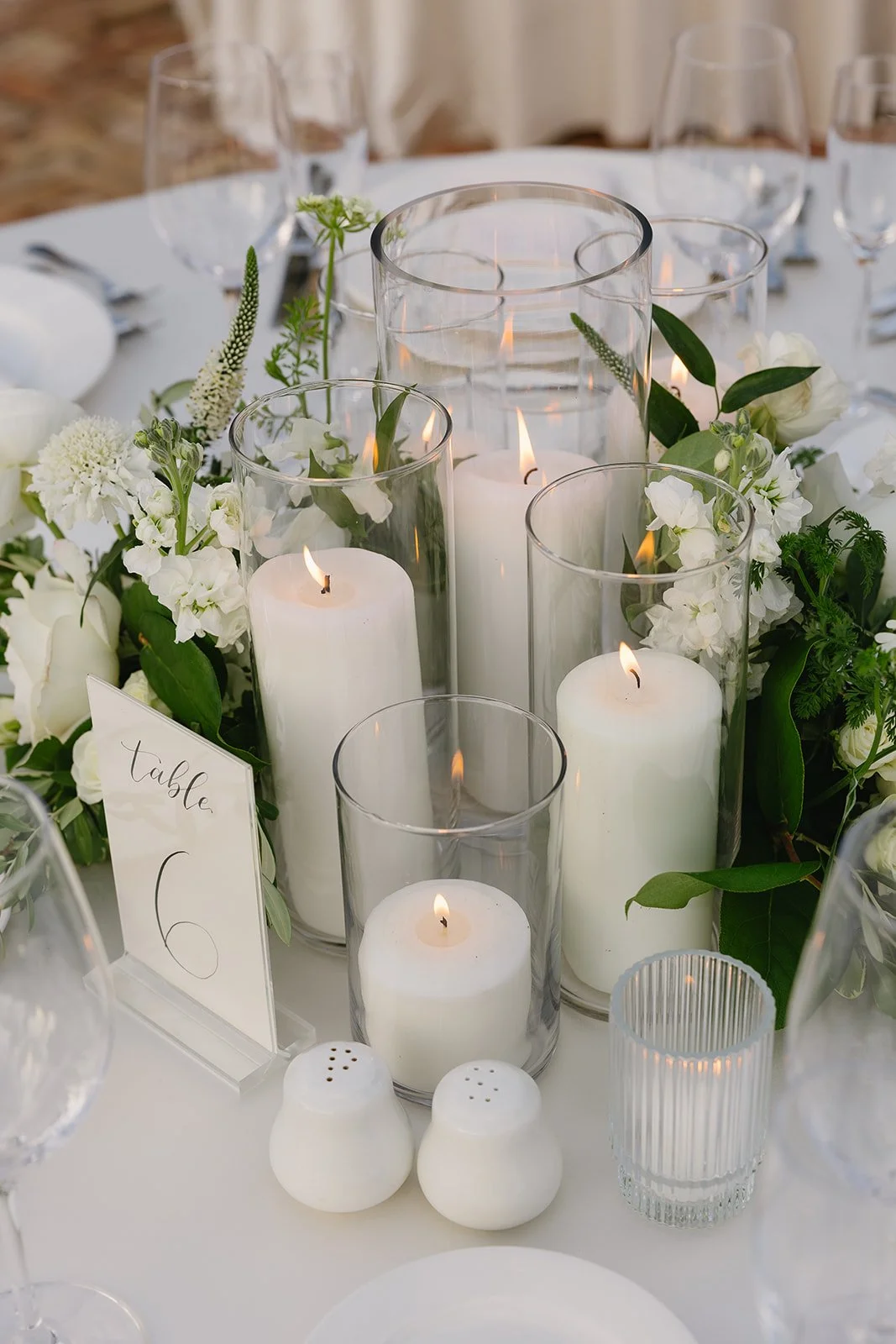 Table centerpiece with white candles in glass holders, surrounded by white flowers and green foliage, with a table number card and glassware visible.