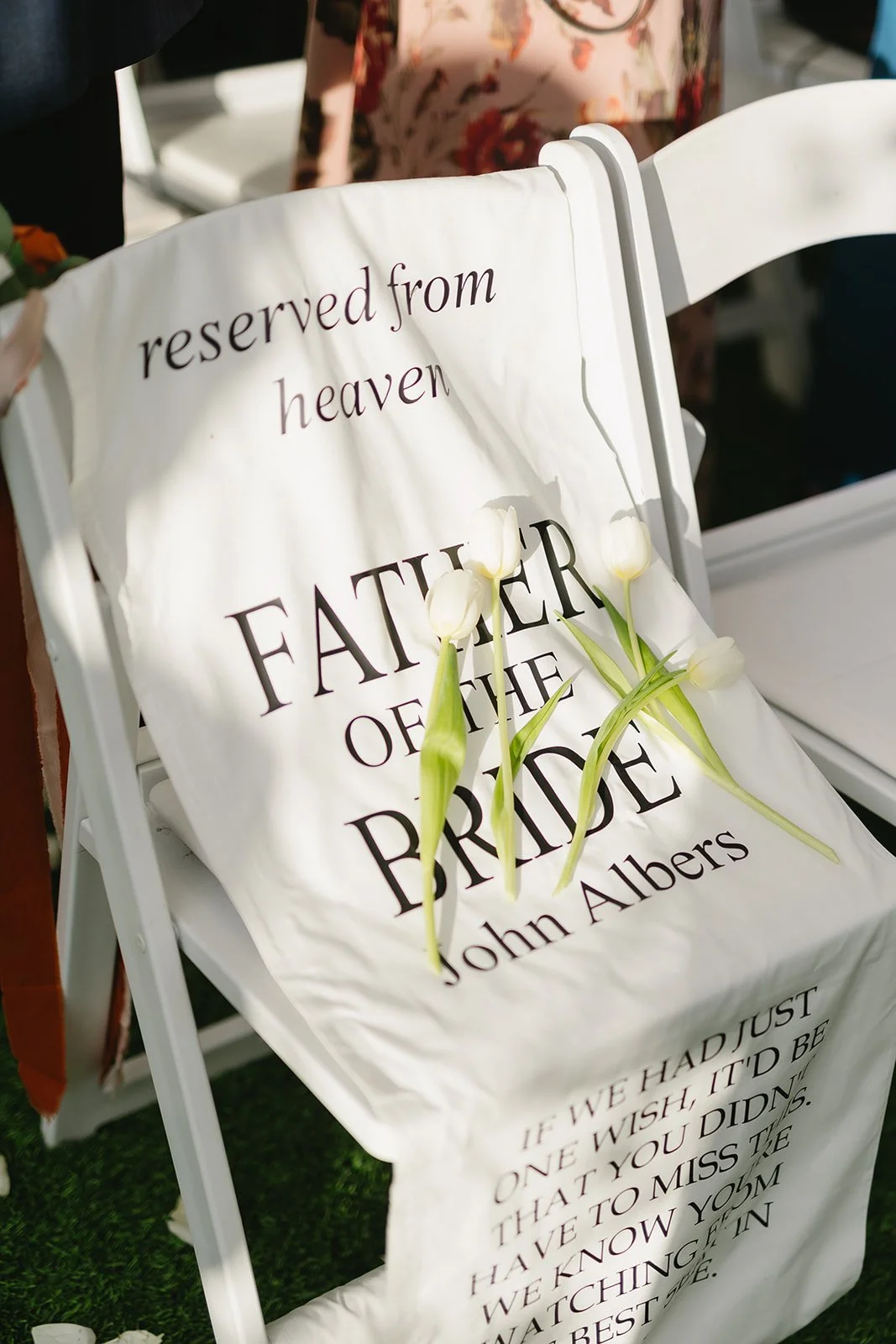A white chair with a fabric sign draped over it that reads 'reserved from heaven, FATHER OF THE BRIDE, John Albers'. The sign has white tulips placed on it. The background shows people at a wedding or event celebration.
